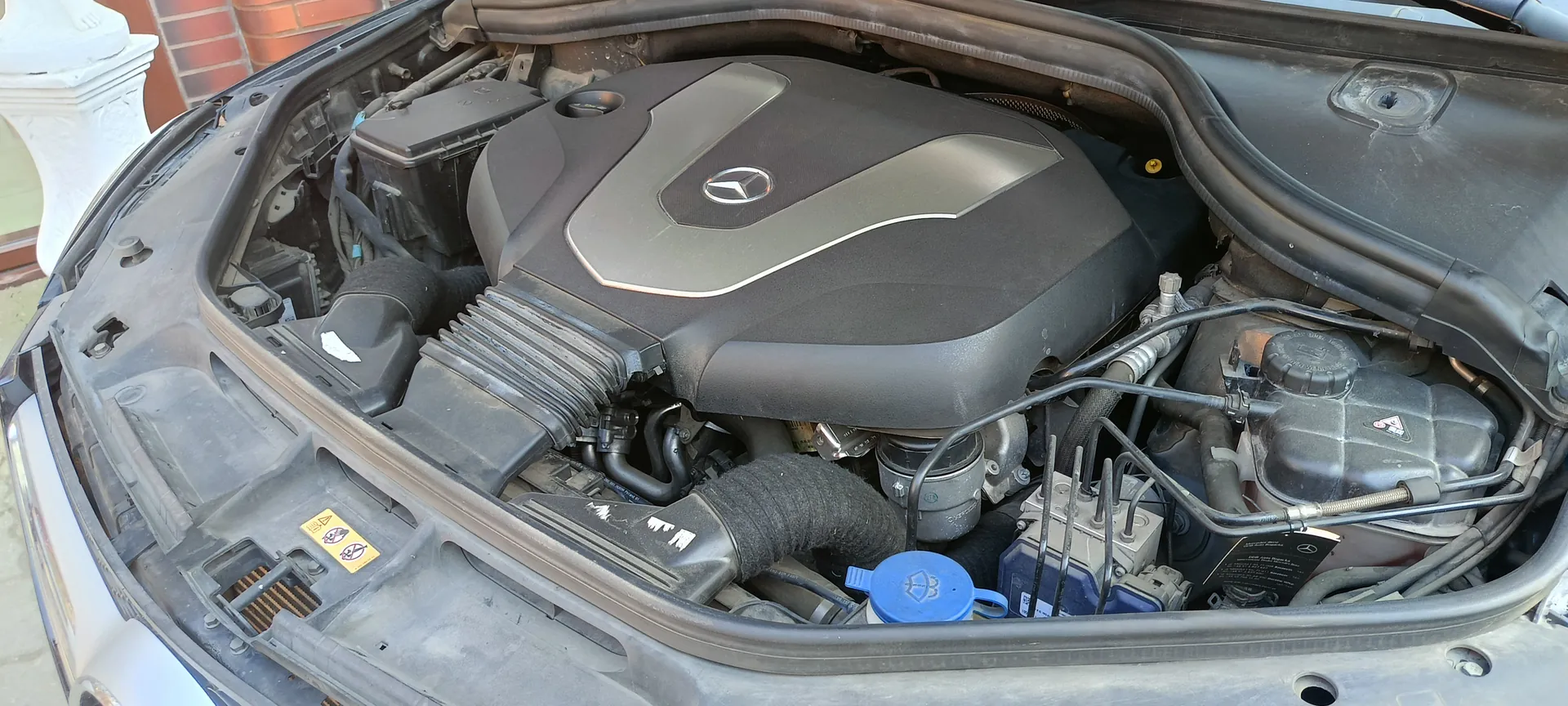 Otwarta kapota samochodu marki Mercedes-Benz z widocznym silnikiem. Na pokrywie silnika znajduje się logo Mercedes-Benz. W silniku widoczne są różne elementy, w tym niebieska pokrywka z symbolem, żółta tabliczka ostrzegawcza z symbolami oraz przewody i rury. W tle ceglana ściana i biała konstrukcja.