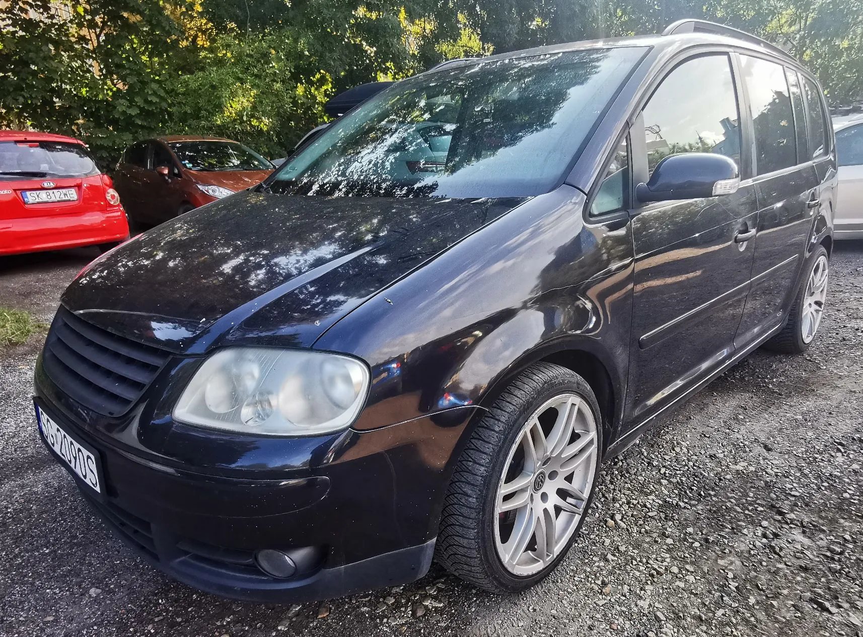 Czarny samochód typu minivan zaparkowany na żwirowym podłożu. Widoczny jest przód i prawy bok pojazdu z dużymi, srebrnymi felgami aluminiowymi. W tle widać czerwony samochód z tablicą rejestracyjną SK 812WE oraz brązowy samochód. W tle znajdują się zielone drzewa.