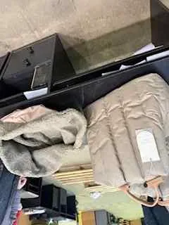 Wnętrze sklepu z beżową pikowaną kurtką na czarnym stole. Na kurtce leży szara tkanina lub koc. W tle widoczne półki z różnymi przedmiotami oraz czarna szafka z szufladami. Podłoga z betonu.