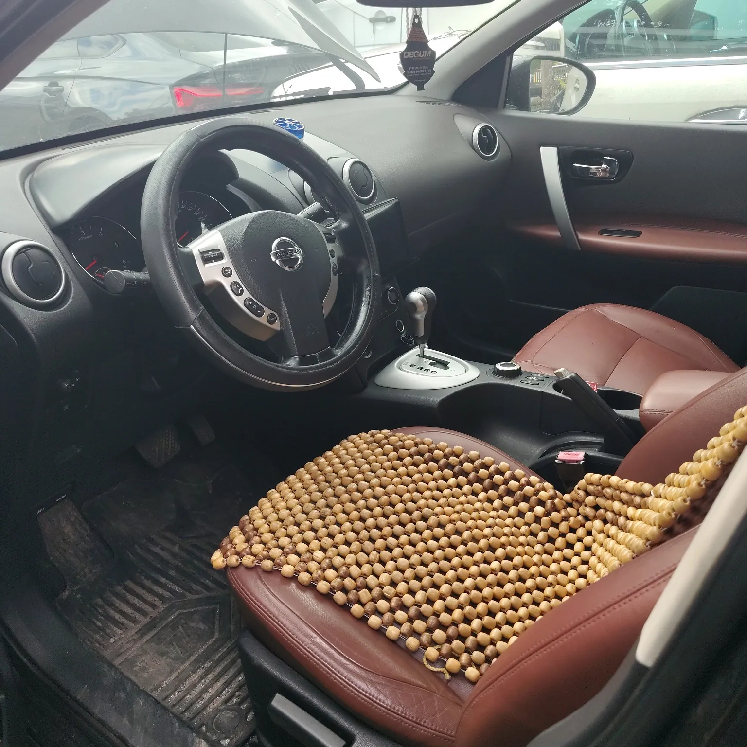 Wnętrze samochodu marki Nissan z perspektywy kierowcy. Widoczna jest czarna kierownica z logo Nissana, deska rozdzielcza, lewy fotel w kolorze brązowym, na którym leży beżowa nakładka z drewnianych koralików. W tle widać fragmenty innych samochodów za szybą przednią.