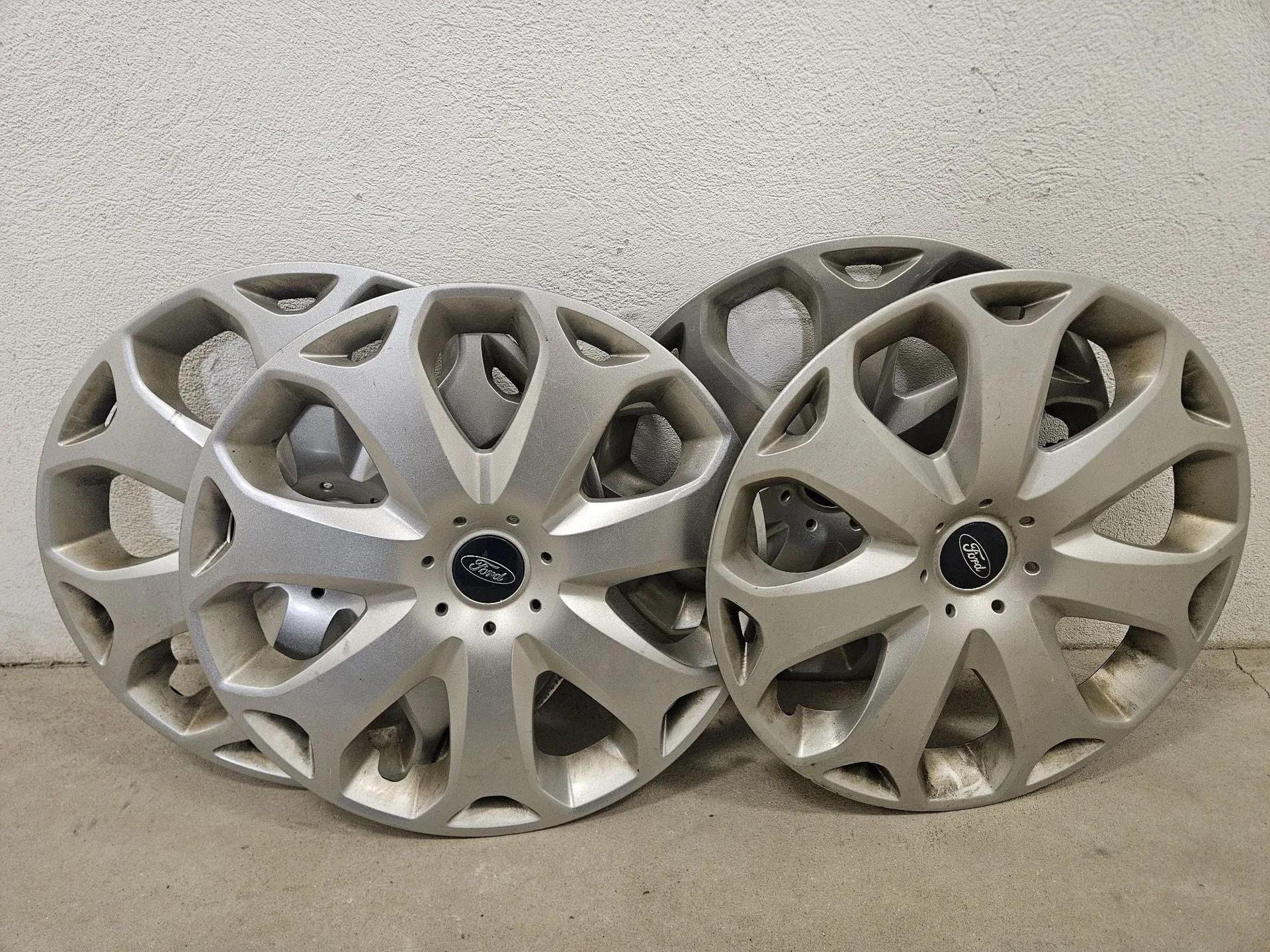 Cztery srebrne, plastikowe kołpaki do kół samochodowych marki Ford, o wieloramiennym wzorze, oparte o jasną, chropowatą ścianę. Na środku każdego kołpaka znajduje się ciemne logo Ford.