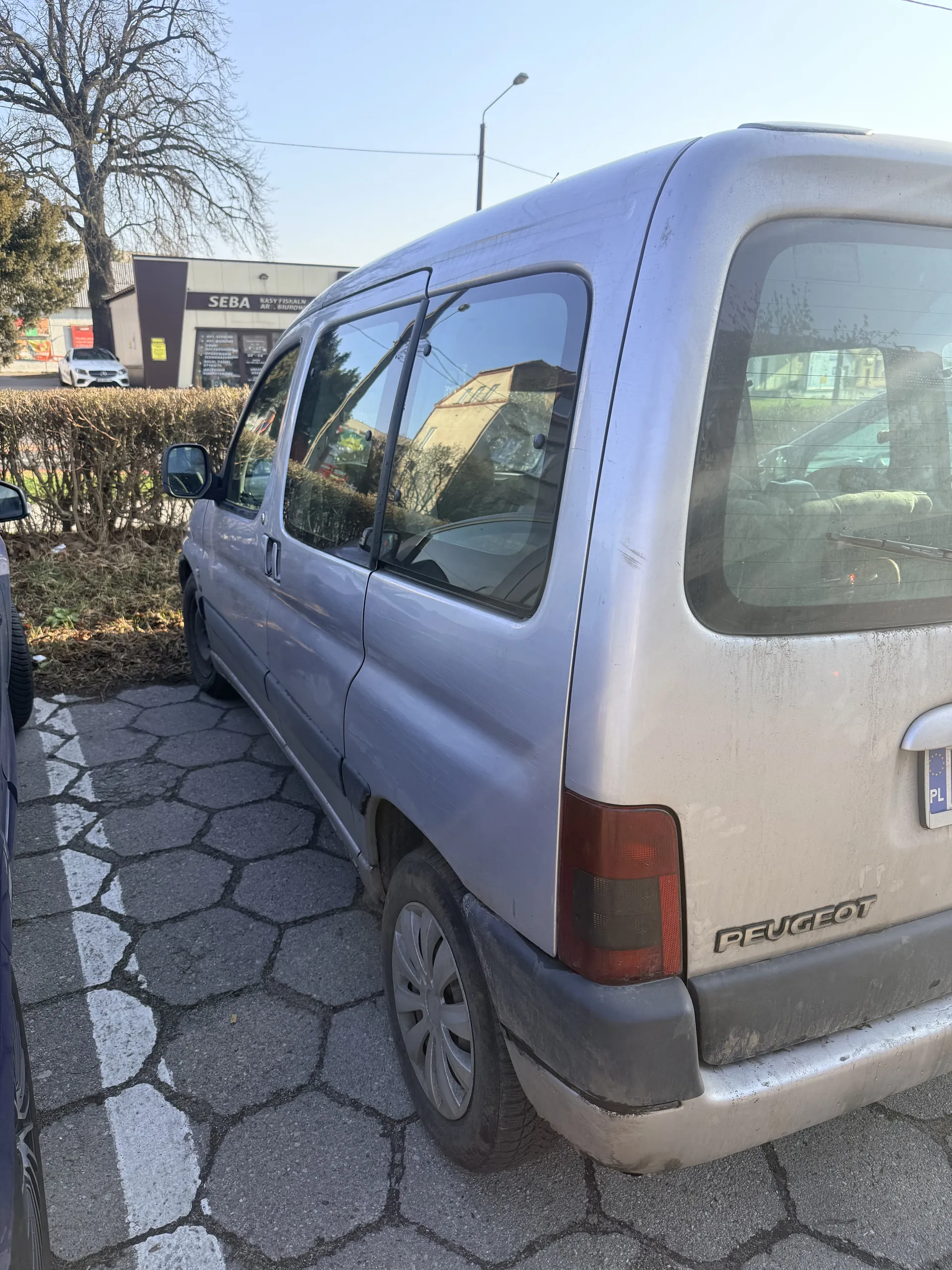 Srebrny samochód marki Peugeot zaparkowany na brukowanej powierzchni z sześciokątnymi płytami. Widoczna tylna część pojazdu z napisem 'PEUGEOT' oraz tablicą rejestracyjną. W tle budynek z napisem 'SEBA', drzewa bez liści i inne pojazdy. Odbicia w szybach przedstawiają otoczenie. Samochód ma ślady brudu na nadwoziu.