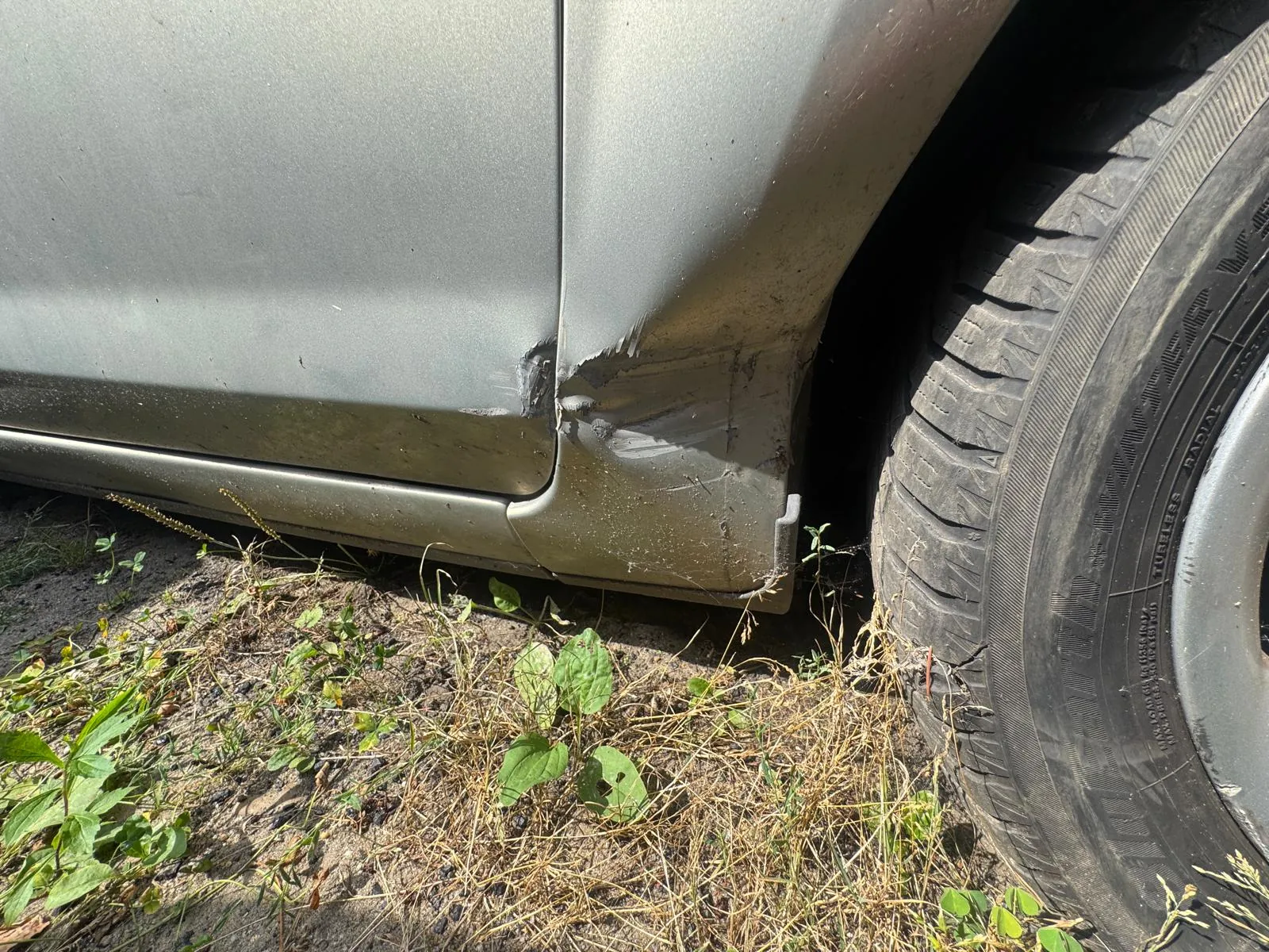 Zbliżenie na uszkodzony element nadwozia srebrnego samochodu w okolicy przedniego nadkola. Blacha jest wgnieciona i porysowana. Obok widoczna jest opona samochodu oraz fragment podłoża z ziemią i suchą trawą.