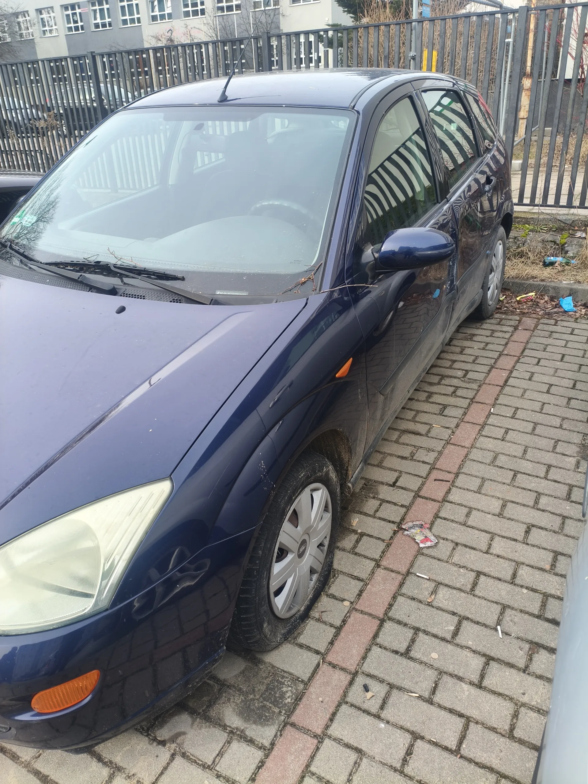 Ciemnoniebieski samochód osobowy typu hatchback zaparkowany na brukowanej powierzchni. Widoczne są przednie światła, lusterko boczne, felgi oraz pomarańczowe oznaczenia. W tle metalowe ogrodzenie, budynki z oknami oraz rozrzucone śmieci na ziemi. Samochód znajduje się pod kątem, z przodem skierowanym w lewo. Na przedniej szybie widoczne są drobne gałązki.