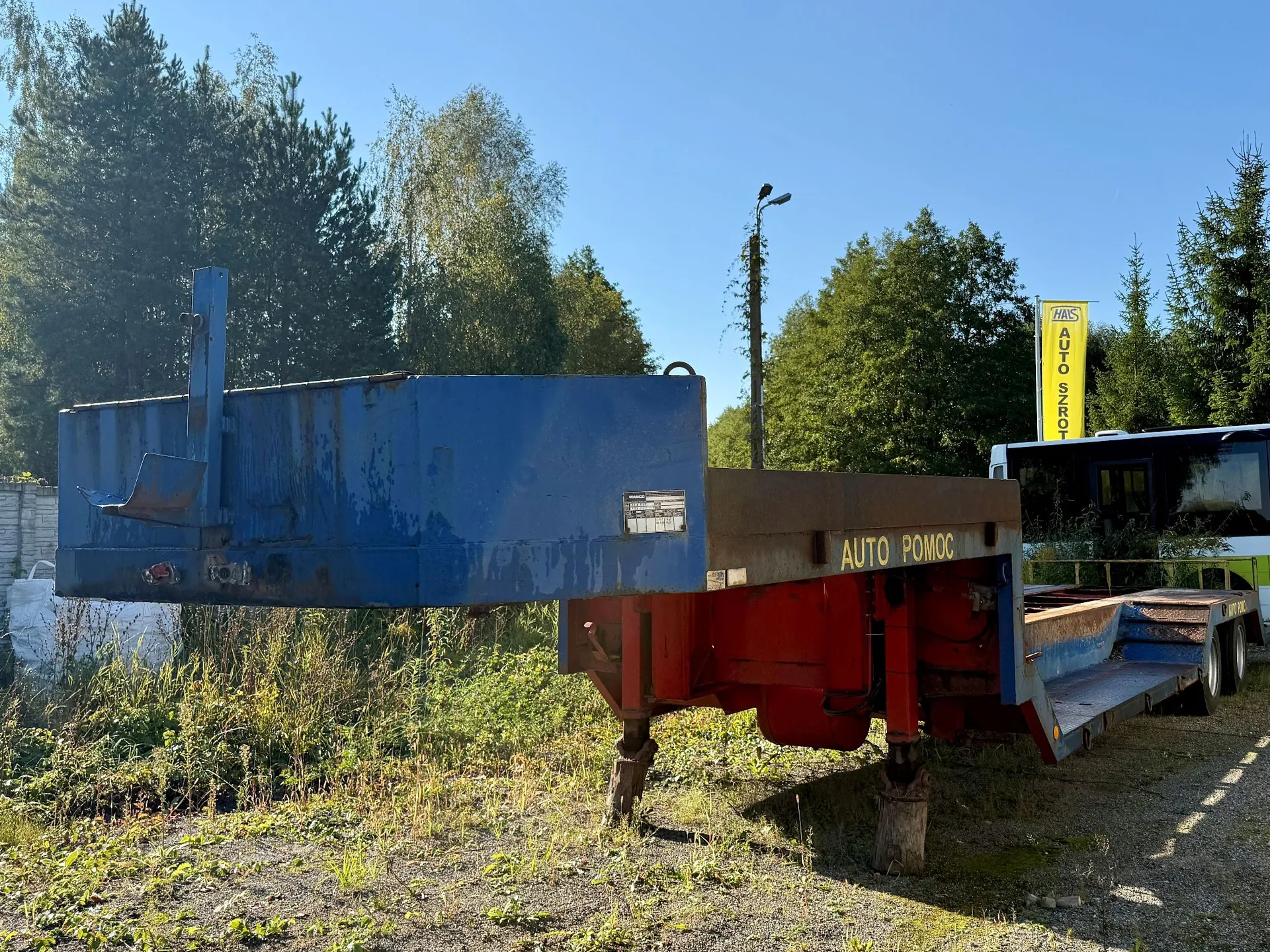 Zdjęcie przedstawia lawetę samochodową zaparkowaną na trawiastym terenie. Przednia część lawety jest niebieska i podniesiona do góry, tylna część jest czerwona z napisem 'AUTO POMOC'. W tle widoczne są zielone drzewa, niebieskie niebo oraz fragment białego pojazdu i żółty pionowy znak z napisem 'AUTO ZŁOM'.