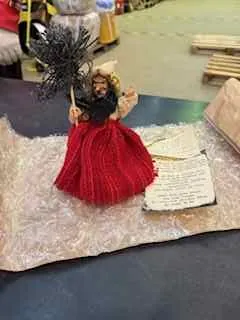 Lalka w czerwonej sukience z czarnymi włosami trzymająca miotłę. W tle widoczne są otwarte książki z nieczytelnym tekstem, pudełko oraz stos patyków. Obiekty umieszczone na powierzchni z jasnym wzorem.