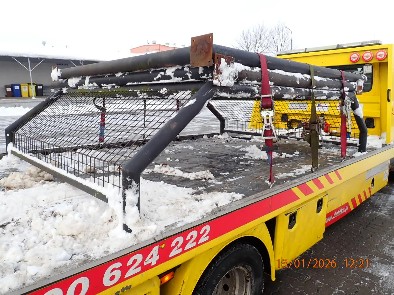 Żółty wóz holowniczy z czerwonym pasem i białymi cyframi 624 222 oraz adresem www.dahika.pl na boku. Na płycie załadunkowej znajdują się metalowe rury przywiązane czerwonymi paskami, częściowo pokryte śniegiem. W tle widoczne są budynki, pojemniki na śmieci oraz drzewa bez liści. Na obrazie widnieje data i godzina: 13/01/2026 12:21.
