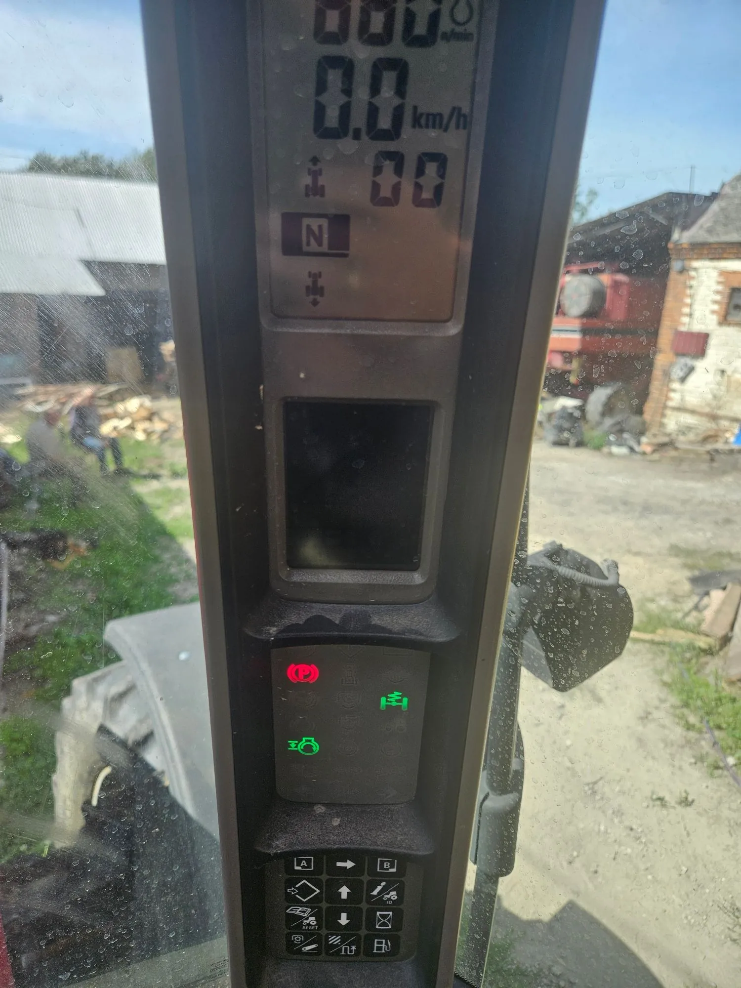 Zbliżenie na panel sterowania i wyświetlacz wewnątrz kabiny ciągnika. Na górnym cyfrowym wyświetlaczu widnieje prędkość 0.0 km/h, litera N oznaczająca bieg jałowy oraz inne wskaźniki techniczne. Poniżej znajduje się panel z kontrolkami: czerwoną ikoną hamulca ręcznego oraz dwiema zielonymi ikonami. Na samym dole znajduje się panel z przyciskami funkcyjnymi z różnymi symbolami, takimi jak strzałki, ikona samochodu i inne oznaczenia techniczne.