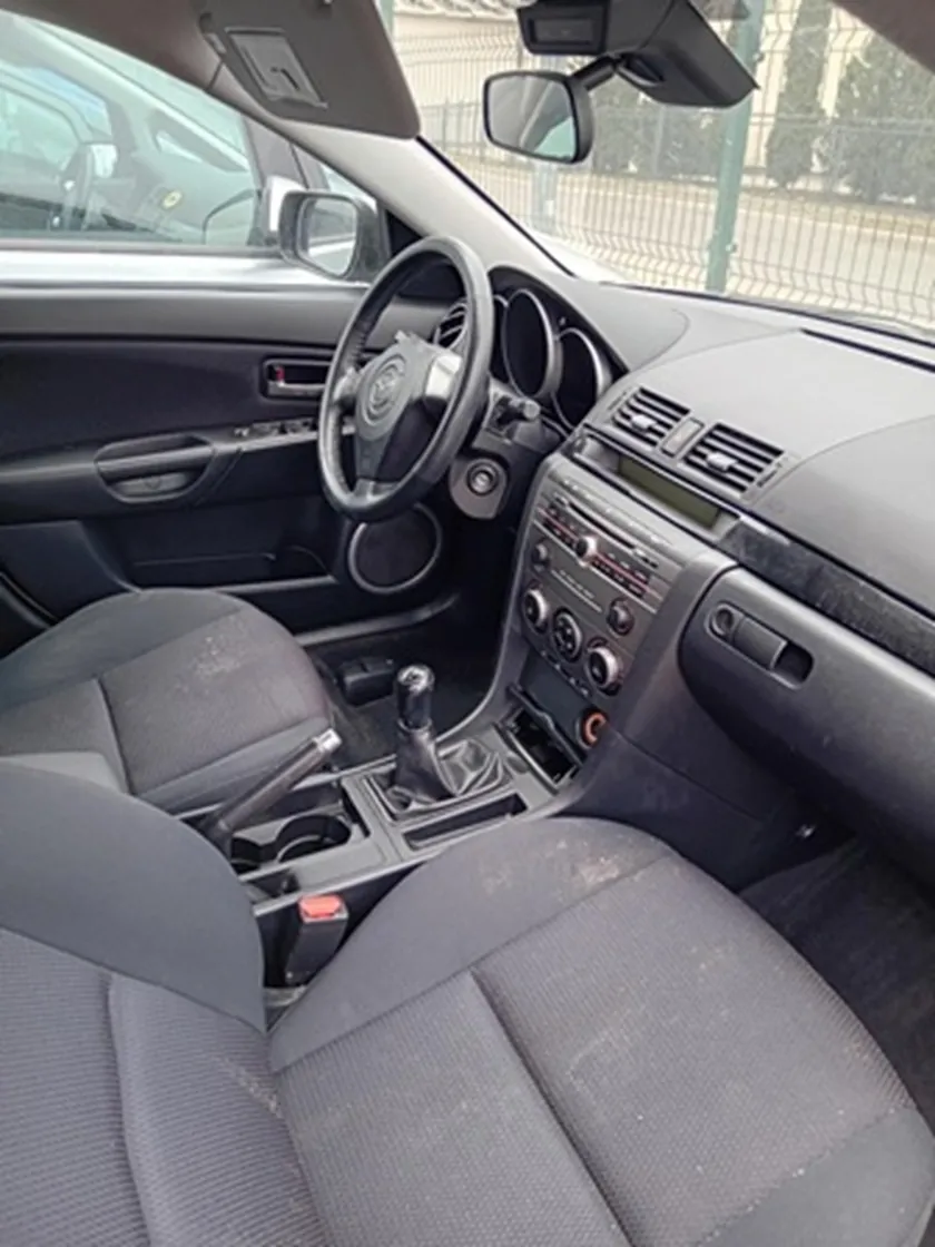 Wnętrze samochodu z widocznymi szarymi tkaninowymi fotelami, kierownicą po lewej stronie, deska rozdzielcza z wentylacjami, radio z przyciskami i pokrętłami, dźwignią zmiany biegów w centralnej konsoli. Przez przednią szybę widoczny płot i budynki na zewnątrz. Wnętrze wydaje się nieco zabrudzone.