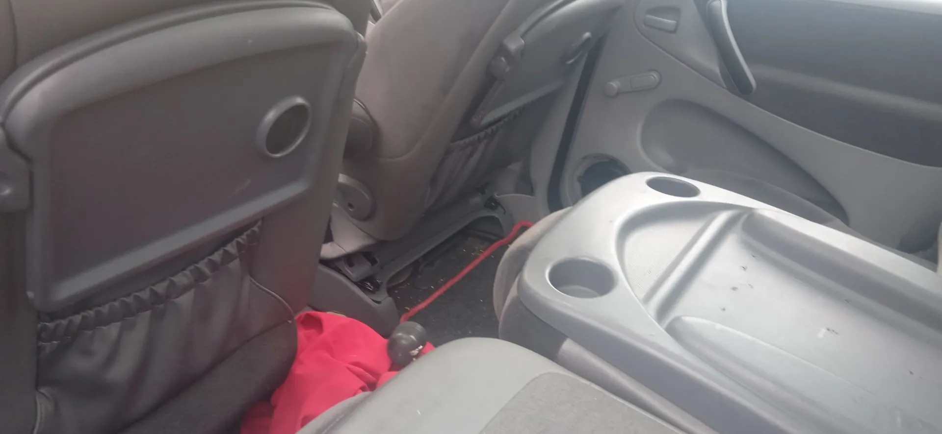 Wnętrze samochodu pokazane z perspektywy tylnego siedzenia. Po lewej stronie widoczny jest oparcie fotela z kieszenią i uchwytem na kubek. Po prawej stronie znajduje się plastikowy podłokietnik z dwoma uchwytami na napoje. W tle widoczny jest fragment drzwi pasażera oraz czerwona tkanina leżąca na podłodze pojazdu.