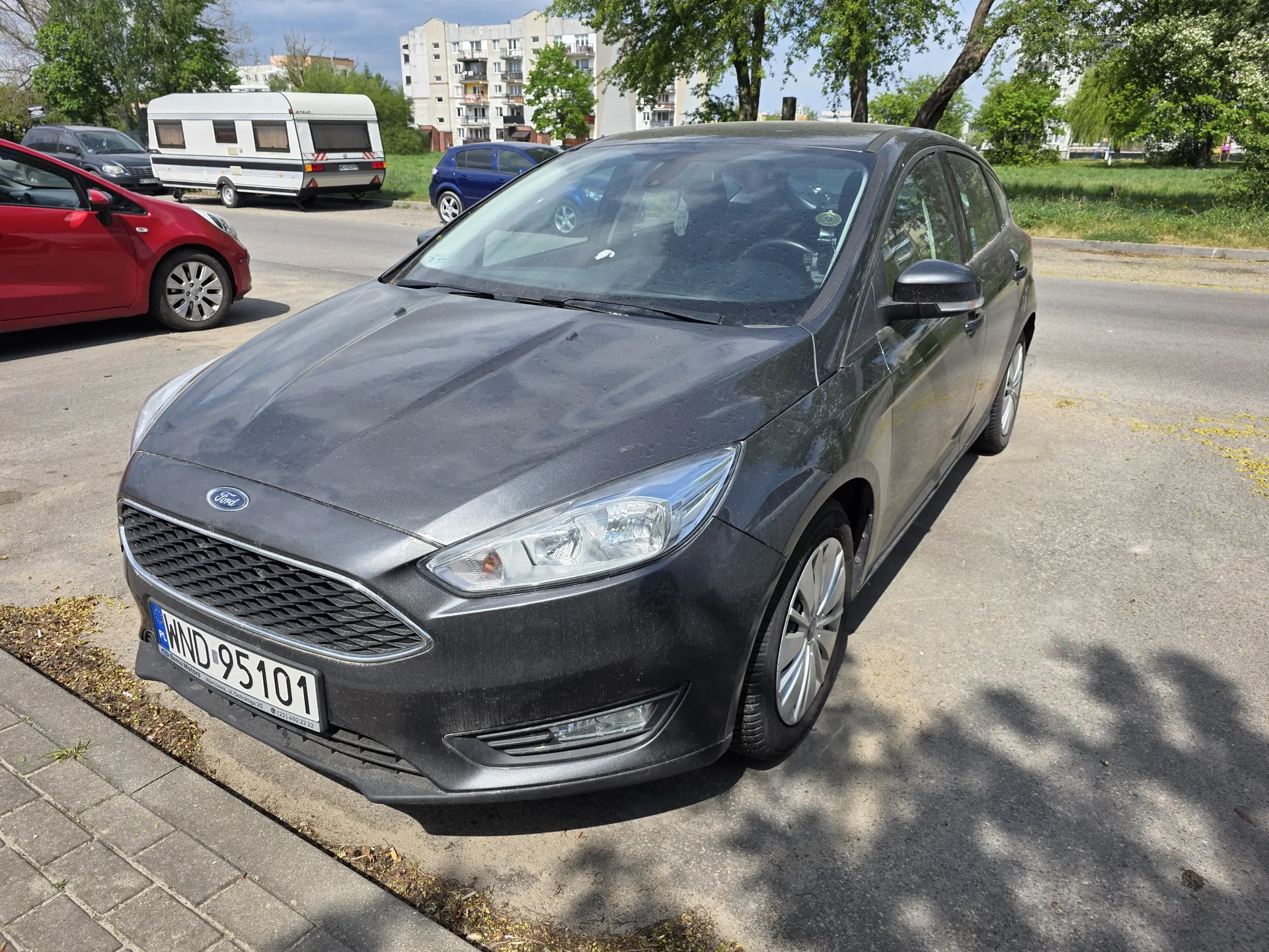 Czarny samochód marki Ford z tablicą rejestracyjną WND 95101 zaparkowany na asfaltowym placu. W tle widoczne są inne pojazdy: czerwony samochód, niebieski samochód i biały przyczepa. Za nimi stoją bloki mieszkalne, drzewa oraz trawnik. Na pierwszym planie fragment chodnika z bruku i suchych liści.