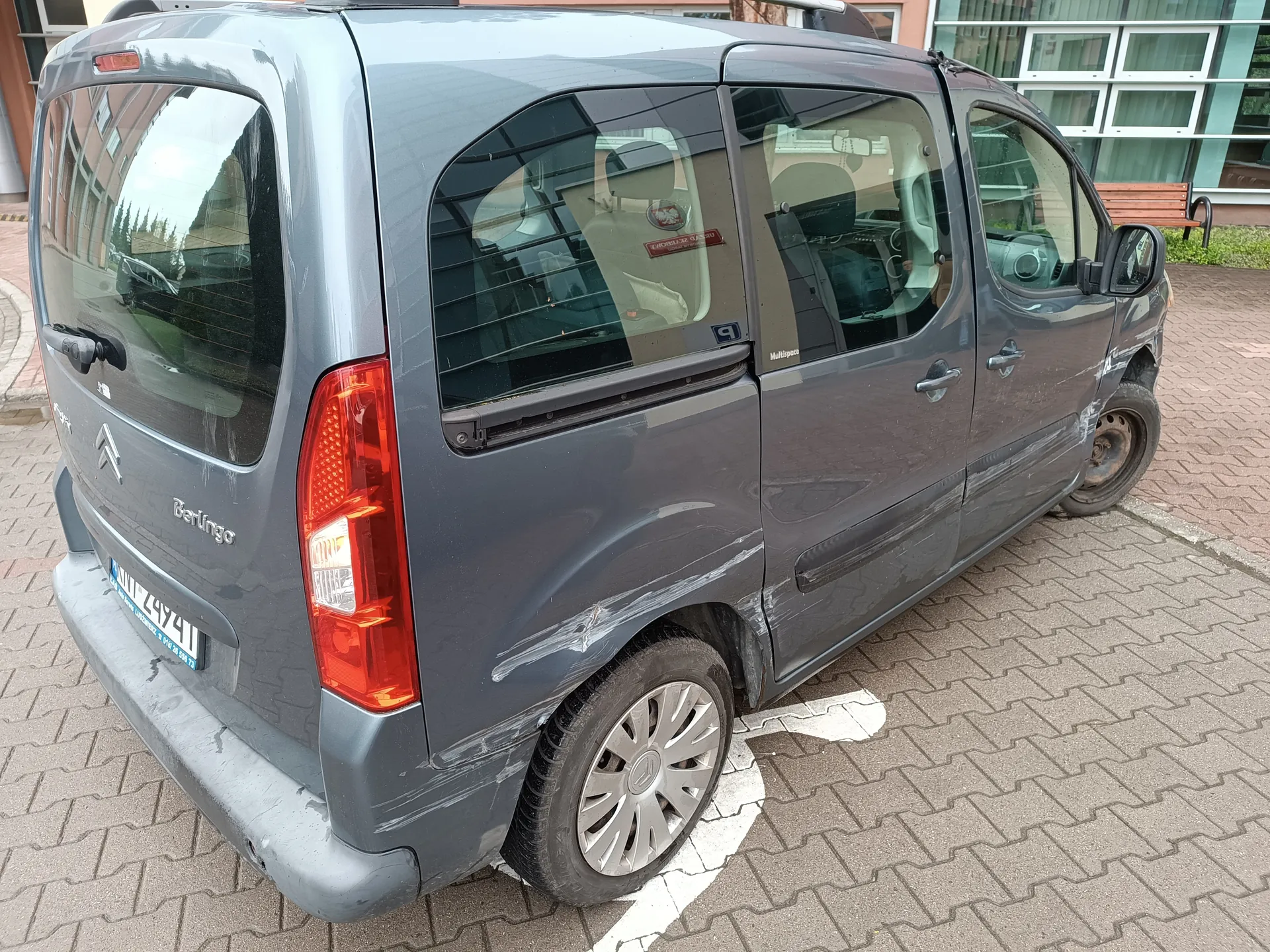Szary samochód Citroën Berlingo zaparkowany na brukowanej powierzchni. Widoczne są uszkodzenia na boku pojazdu, w tym zadrapania i ślady naprawy. Na tylnym szybie napis "Berlingo" oraz logo marki. Tablica rejestracyjna z nieczytelnym numerem. W tle budynek z dużymi oknami i ławką. Wewnątrz samochodu widoczne siedzenia oraz czerwony napis na tylnym oknie.