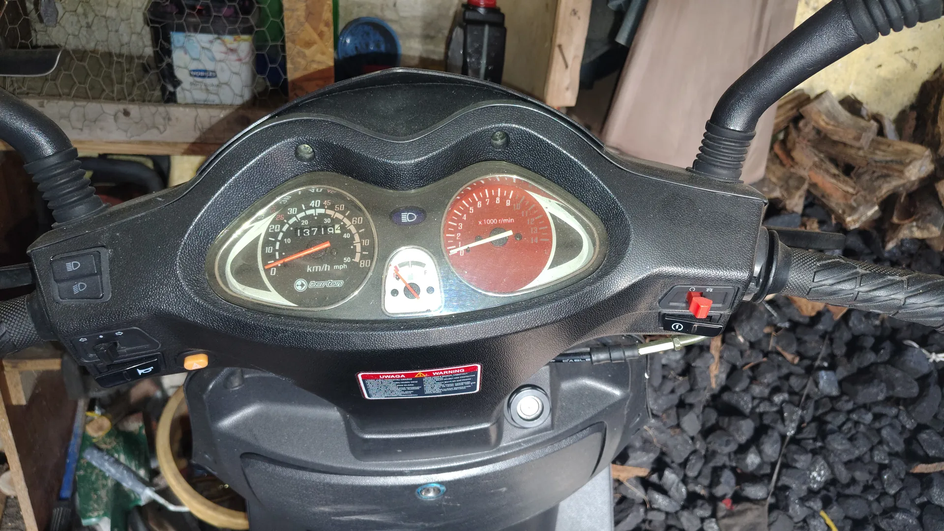 Zbliżenie na czarny deski rozdzielczej skutera z dwoma głównymi wskaźnikami: po lewej prędkościomierz z zakresem do 80 km/h i mph, cyfrą 13719 oraz napisem 'Bajaj', po prawej czerwony tachometr z zakresem do 14 x1000 r/min. W centrum mały wskaźnik paliwa. Poniżej czerwono-biały naklejony napis 'UWAGA' i 'WARNING'. Po bokach widoczne czarne kierownice z przyciskami, w tle narzędzia, siatka, pojemnik i węgiel drzewny na podłodze.