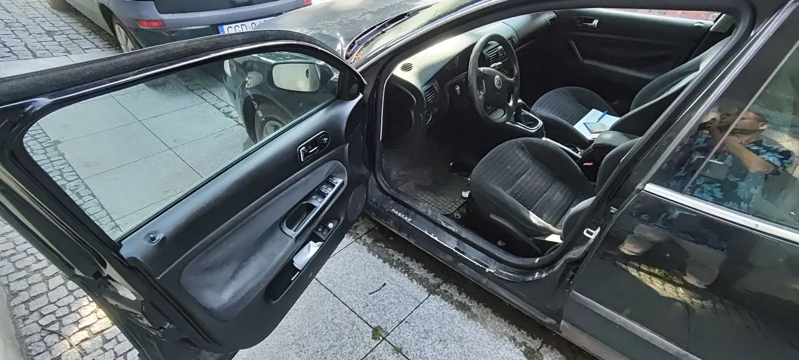 Widok z góry na otwarte drzwi kierowcy czarnego samochodu marki Volkswagen Passat. Wnętrze pojazdu zawiera ciemną tapicerkę, kierownicę z logo Volkswagena oraz fotele. Na progu samochodu widoczny jest napis 'PASSAT'. Auto zaparkowane jest na chodniku z jasnych płyt betonowych i kostki brukowej.