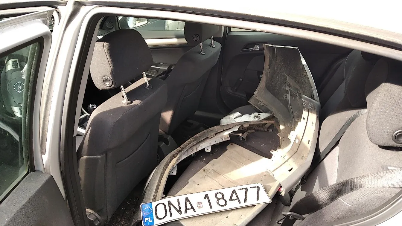Wnętrze samochodu widziane z otwartych drzwi pasażera. Na tylnej kanapie leży duży, jasnoszary element plastikowy, prawdopodobnie fragment zderzaka lub osłony podwozia, do którego przymocowana jest tablica rejestracyjna o numerze ONA 18477 z oznaczeniem PL.