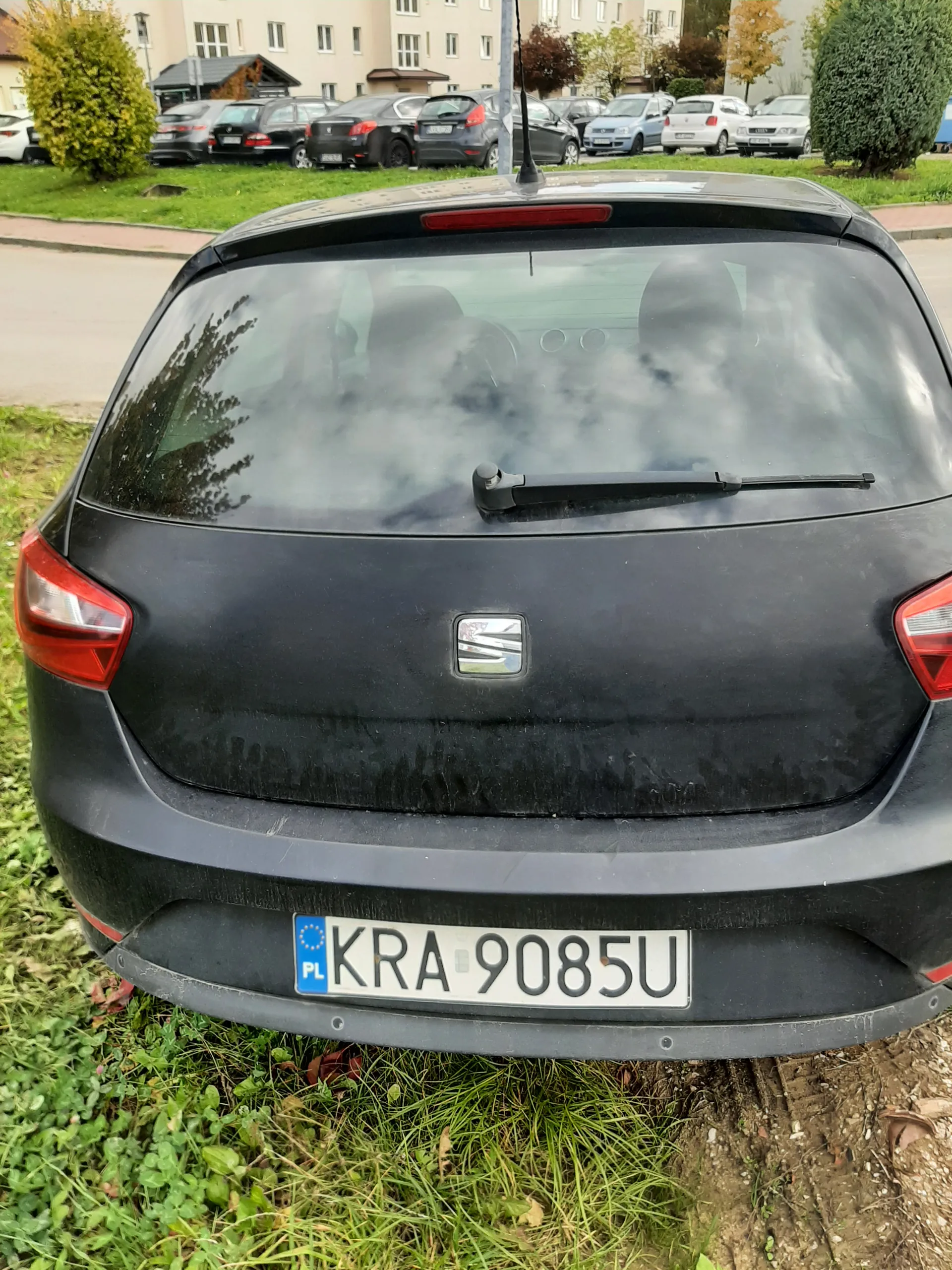 Tył czarnego samochodu marki Honda z tablicą rejestracyjną KRA 9085U. Samochód zaparkowany na trawie obok drogi. W tle widoczne inne zaparkowane samochody, budynki mieszkalne oraz krzewy i drzewa.