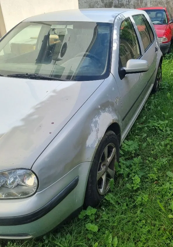 Srebrny samochód osobowy zaparkowany na trawie. Widoczny jest przód i prawy bok pojazdu. Na prawym przednim błotniku widoczne są uszkodzenia lakieru i wgniecenia. W tle widać fragment innego, czerwonego samochodu.