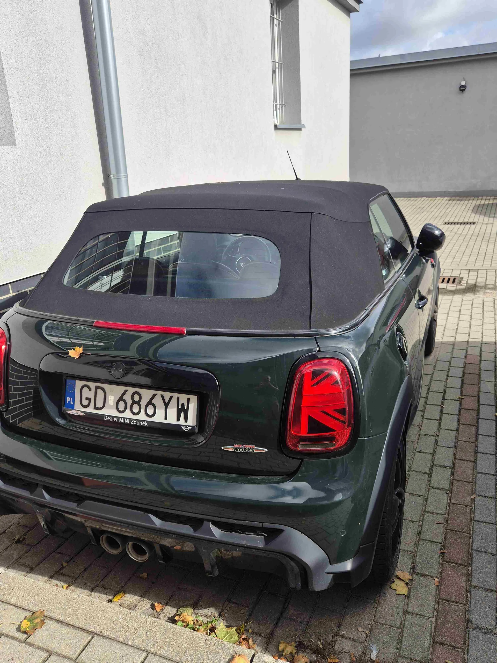 Tylny widok ciemnozielonego samochodu MINI z czarnym, składanym dachem materiałowym. Pojazd posiada polską tablicę rejestracyjną o numerze GD 686YW oraz napis 'Dealer MINI Zdunek' na ramce tablicy. Na klapie bagażnika widnieje emblemat 'John Cooper Works'. Samochód zaparkowany jest na kostce brukowej obok białego budynku.