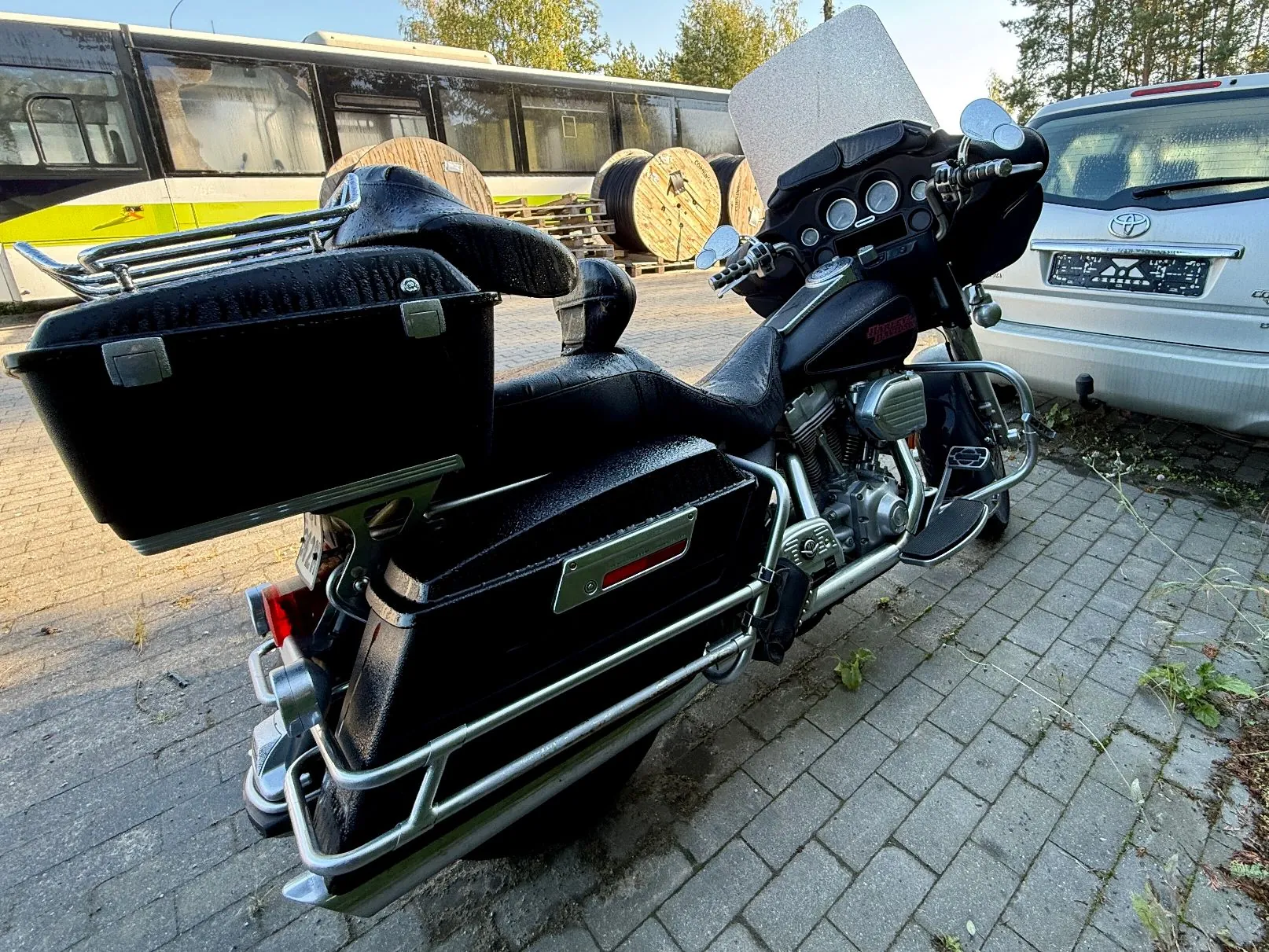 Czarny motocykl typu cruiser z dużymi kuframi bocznymi i tylnym bagażnikiem, zaparkowany na kostce brukowej. W tle widoczny jest żółto-biały autobus oraz srebrny samochód marki Toyota.