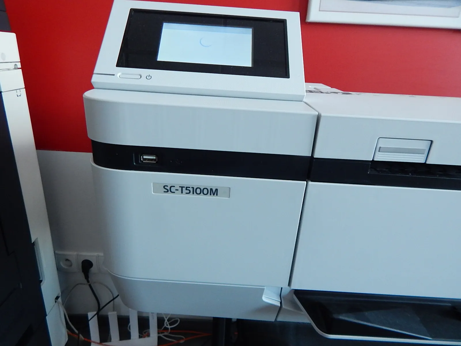Biała drukarka wielkoformatowa z ekranem LCD na górze. Na obudowie urządzenia znajduje się napis 'SC-T5100M'. Urządzenie stoi na tle czerwonej ściany.