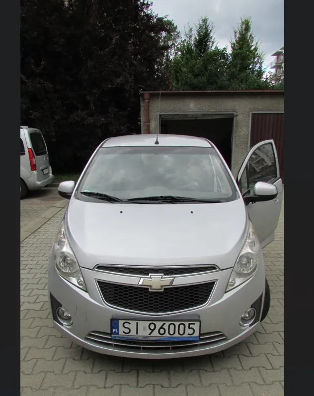Srebrny samochód marki Chevrolet zaparkowany na kostce brukowej przed garażem. Widoczna jest przednia część pojazdu z tablicą rejestracyjną o numerze SI 96005. Po lewej stronie w tle widać fragment białego samochodu, a w tle znajdują się ciemne drzewa i fragment budynku.
