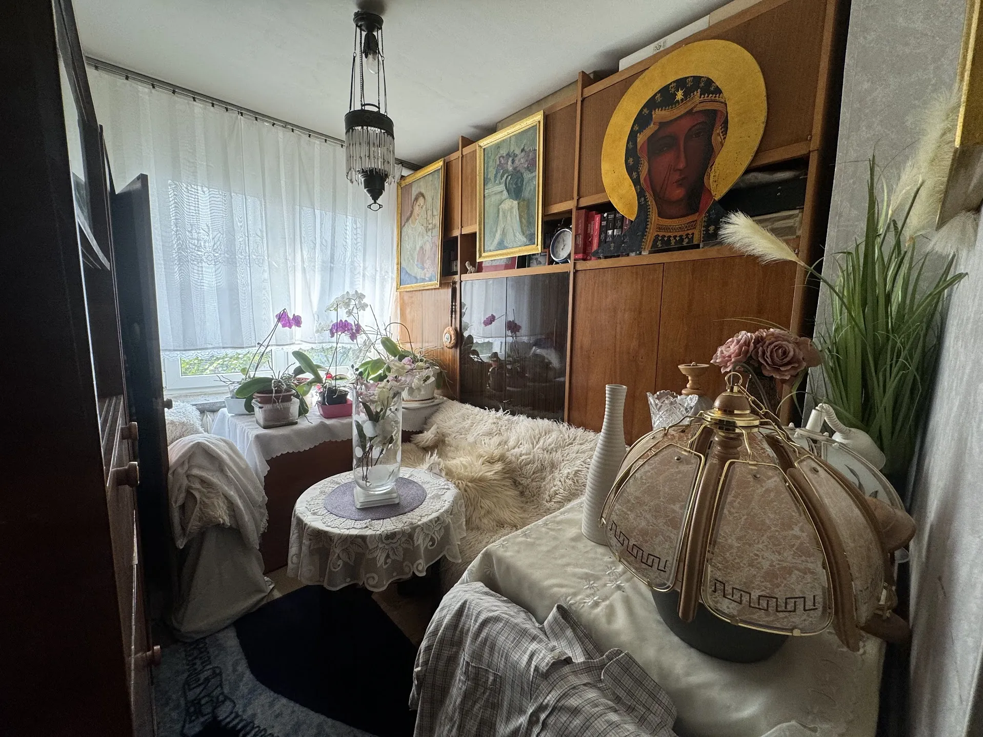 Wnętrze pokoju z drewnianymi szafkami na ścianie, dużym ikoną religijną z złotym nimbem, dwoma ramkowanymi obrazami z kwiatami i postaciami. Na parapecie okna z białą koronką stoją doniczki z fiołkowymi i białymi storczykami. W centrum mały stolik z koronkową serwetką i szklanym wazonem z białymi kwiatami. Pod sufitem wisząca lampa kryształowa. Na podłodze czarny dywan i puszysty biały dywanik. Po prawej stronie stolik z lampą o brązowo-złotym kloszu, białym wazonem, szklanymi naczyniami i sztucznymi różami. Widoczne również zielone rośliny oraz częściowo zakryte meble tkaniną w kratę. Okno zasłonięte przezroczystą zasłoną, za którą widać zielony krajobraz.