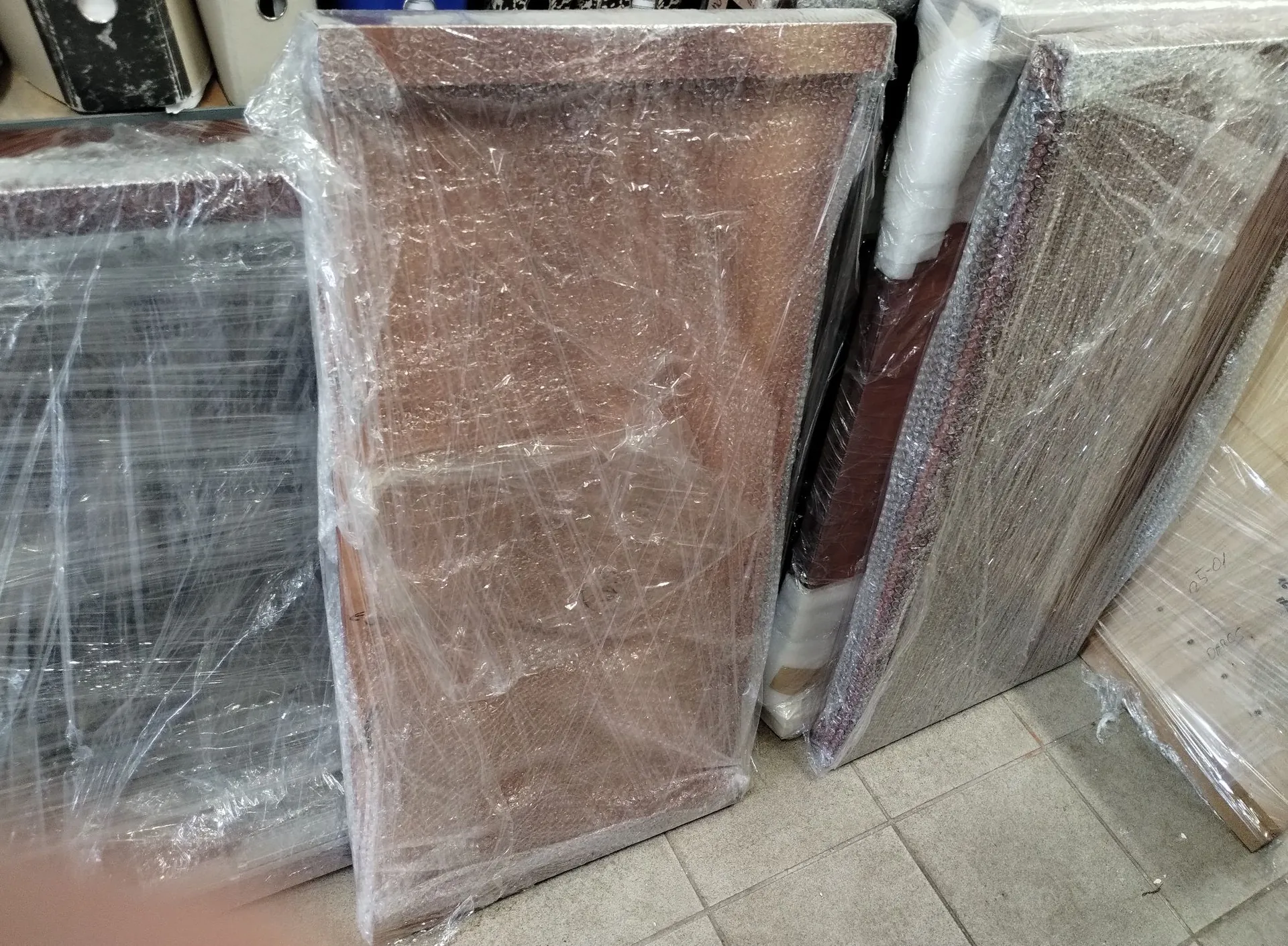 Kilka dużych, płaskich elementów meblowych lub płyt drewnianych, owiniętych w przezroczystą folię stretch, stojących pionowo obok siebie na jasnej, kafelkowej podłodze.