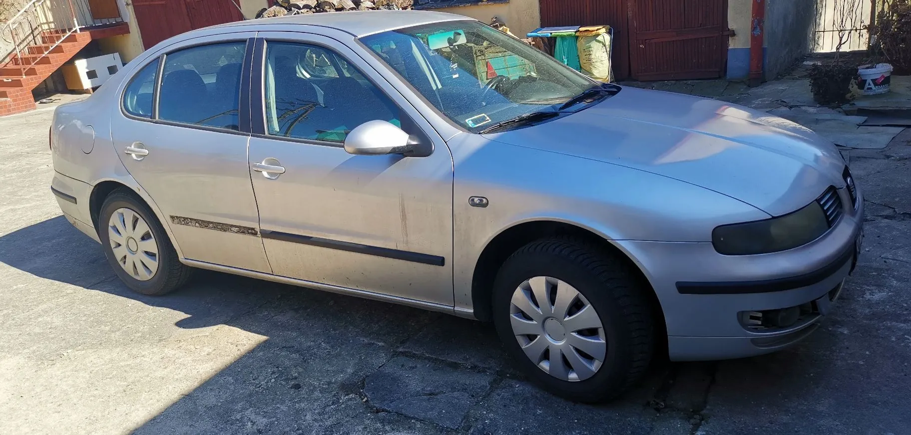Srebrny samochód osobowy marki Volkswagen Passat w nadwoziu sedan, zaparkowany bokiem na betonowym podjeździe. Pojazd ma widoczne ślady zabrudzeń na dolnych partiach nadwozia oraz czarne listwy boczne. W tle widoczny jest fragment budynku z ceglanymi schodami oraz brązowa brama.