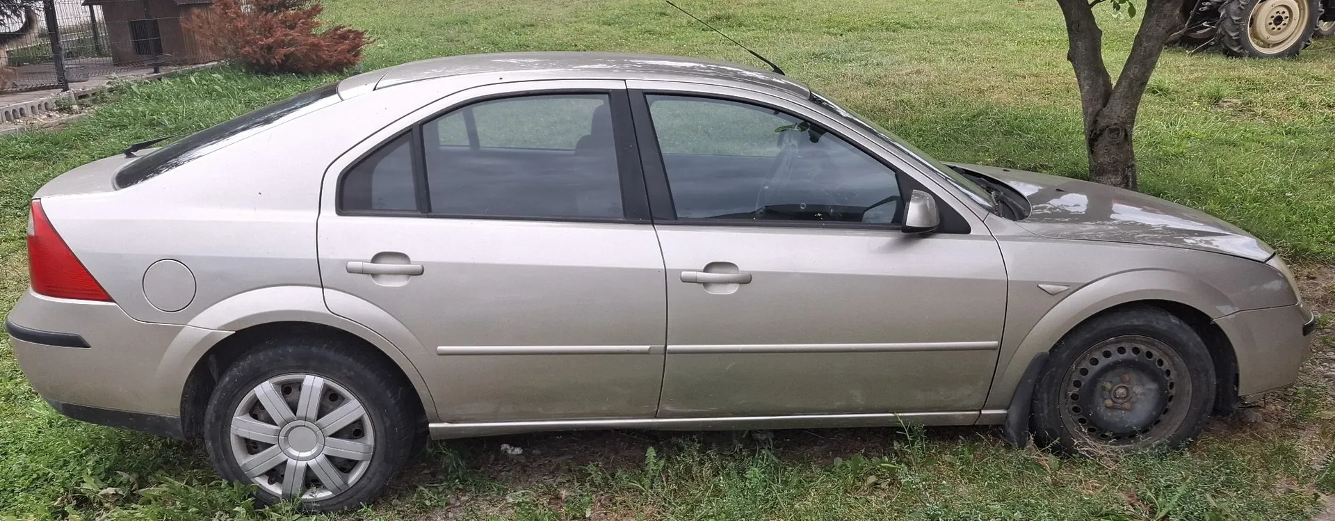 Widok z boku srebrnego samochodu osobowego typu sedan zaparkowanego na trawie. Pojazd ma cztery drzwi, a przednie koło jest stalową felgą bez kołpaka, podczas gdy tylne posiada srebrny kołpak. W tle widoczna jest zielona trawa, drzewo oraz fragment budowli i maszyny rolniczej.