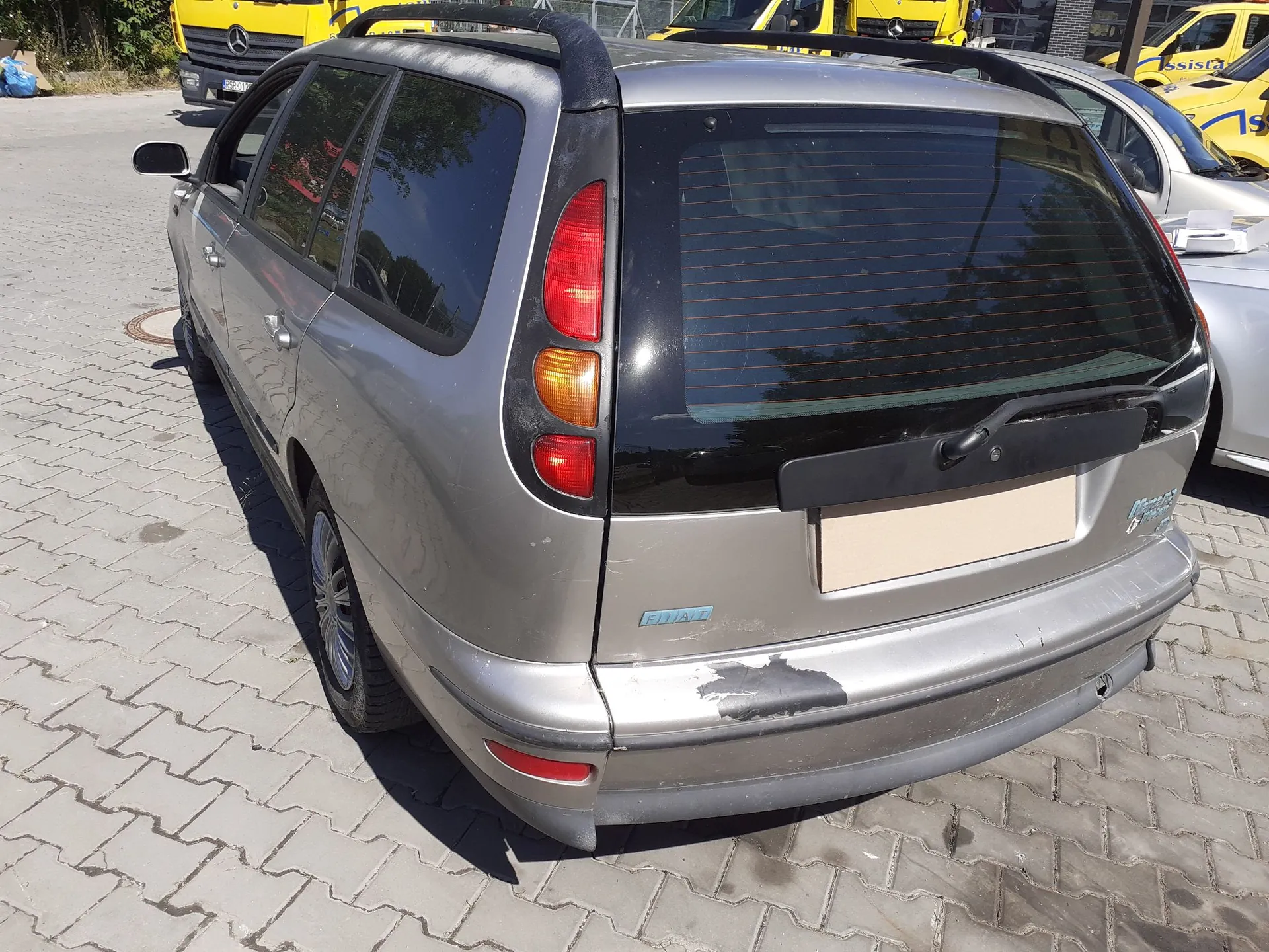 Srebrny samochód marki Fiat w nadwoziu kombi, zaparkowany na kostce brukowej. Zdjęcie przedstawia tył i lewy bok pojazdu. Samochód posiada relingi dachowe, a na tylnym zderzaku widoczne są uszkodzenia lakieru. Tablica rejestracyjna jest zakryta beżowym prostokątem. W tle widoczne są żółte samochody dostawcze.