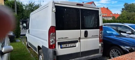 Tylny widok białego vana marki Citroën model Jumper z polską tablicą rejestracyjną o numerze DSW 93101. Pojazd zaparkowany jest obok niebieskiego samochodu osobowego. W tle widoczne są zielone krzewy oraz budynek z czerwonym dachem.