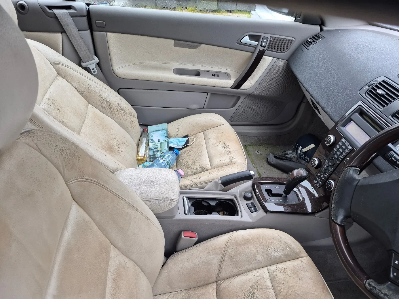 Wnętrze samochodu z brudnymi beżowymi fotelami. Na przednim fotelu pasażera znajdują się plastikowa butelka, niebieski opakunek, maska medyczna i różowy przedmiot. W centralnej konsoli widoczne są uchwyty na napoje, dźwignia zmiany biegów oraz przyciski. Deska rozdzielcza w kolorze szarym z kierownicą po prawej stronie. Na podłodze obok kierowcy leży czarna buty. Drzwi pasażera z beżowym wykończeniem i szarymi elementami, w tym uchwytem i sterami okien.