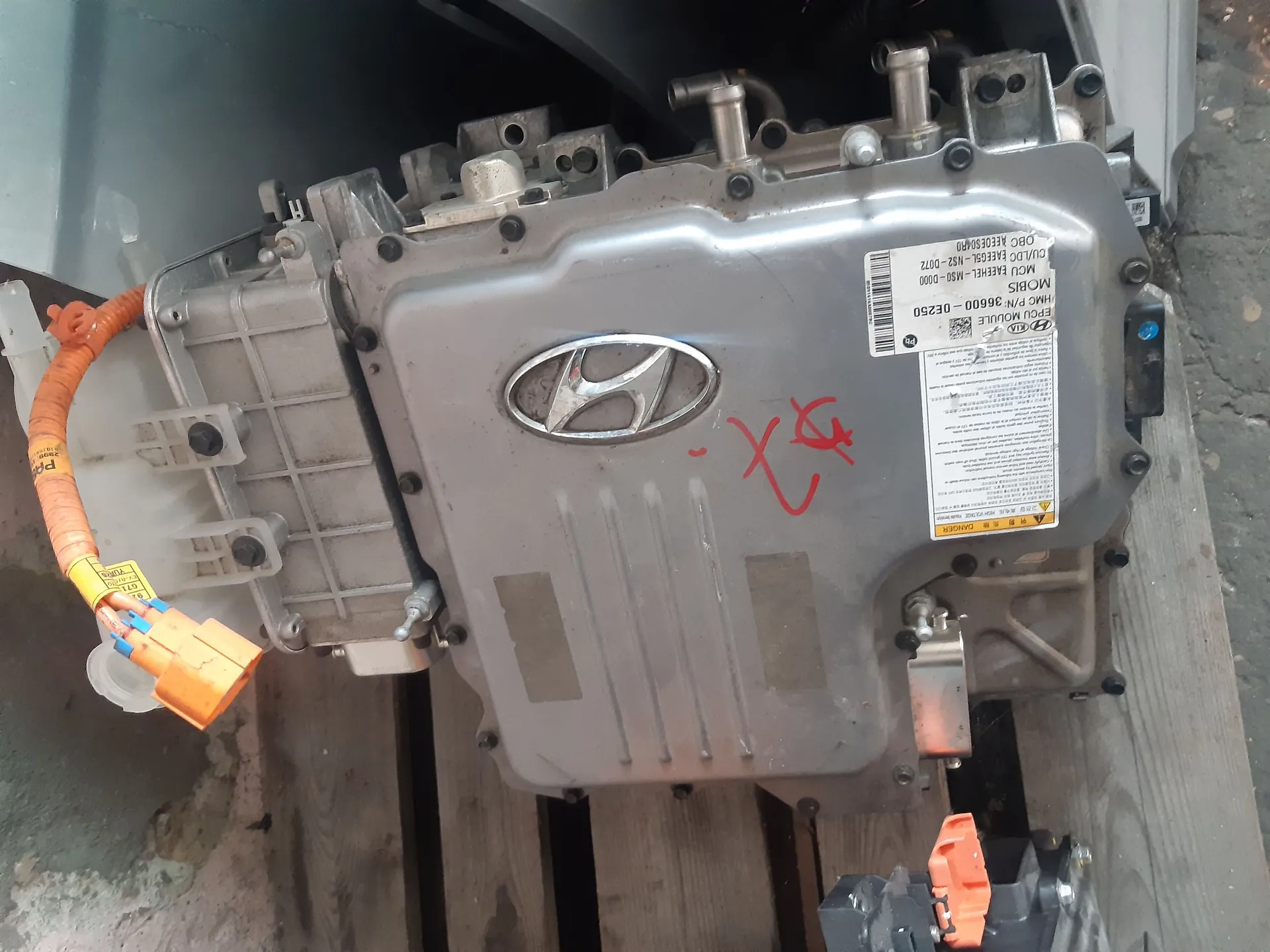 Metalowa obudowa modułu elektronicznego marki Hyundai, prawdopodobnie element układu napędowego pojazdu elektrycznego lub hybrydowego. Na górnej części znajduje się logo Hyundai oraz biała etykieta z danymi technicznymi, w tym napisy: 'MOBIS', 'HMC P/N 36600-DE250' oraz 'CULO/D EO EAEG0L-N22-007E'. Na obudowie widnieje czerwony napis odręczny. Do modułu podłączony jest pomarańczowy przewód wysokiego napięcia z pomarańczową wtyczką. Całość spoczywa na drewnianej palecie.