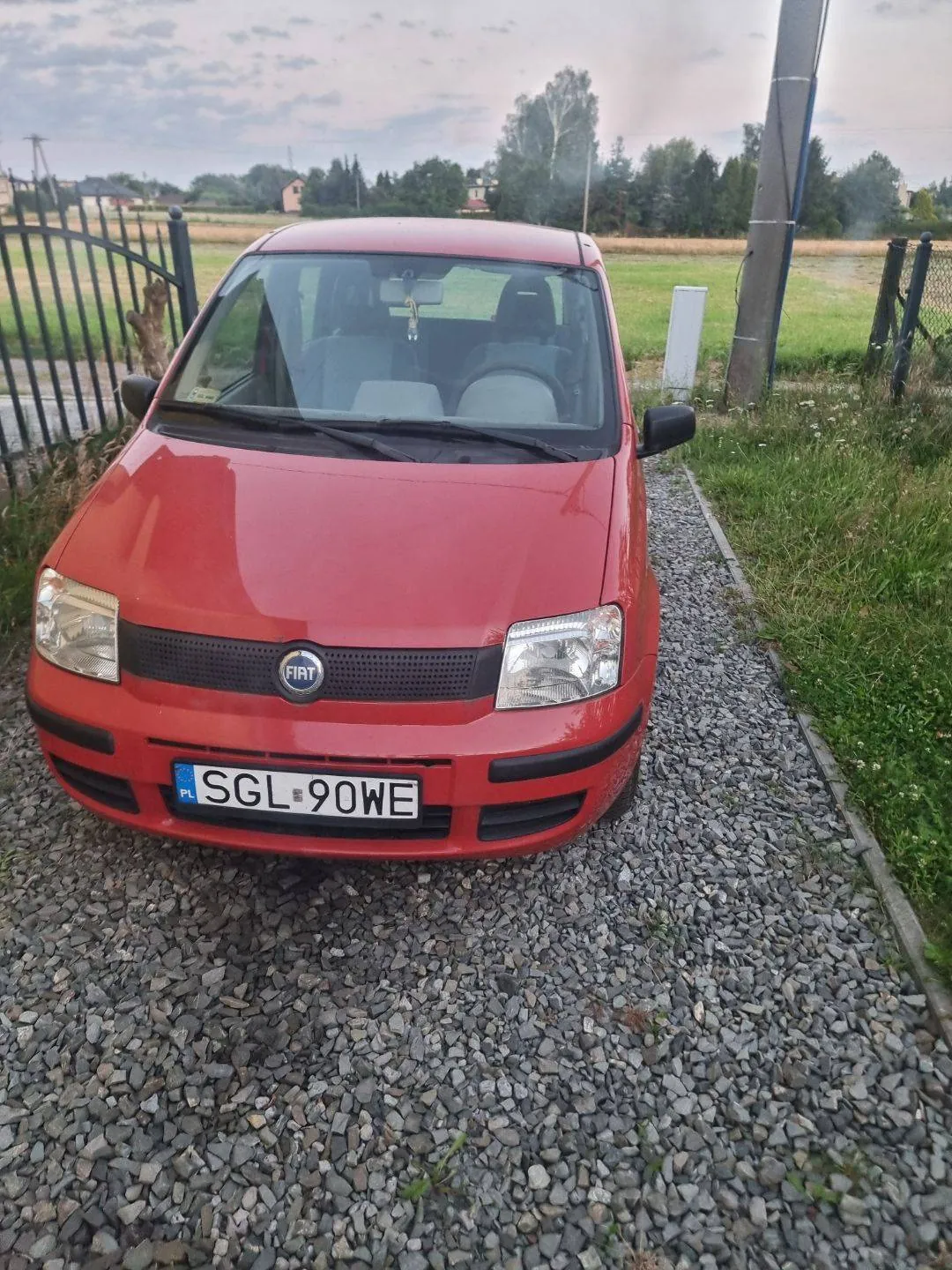 Czerwony samochód marki Fiat zaparkowany przodem na żwirowym podjeździe. Pojazd posiada polską tablicę rejestracyjną o numerze SGL 90WE. W tle widoczny jest metalowy płot, zielona trawa oraz odległe budynki i drzewa pod zachmurzonym niebem.