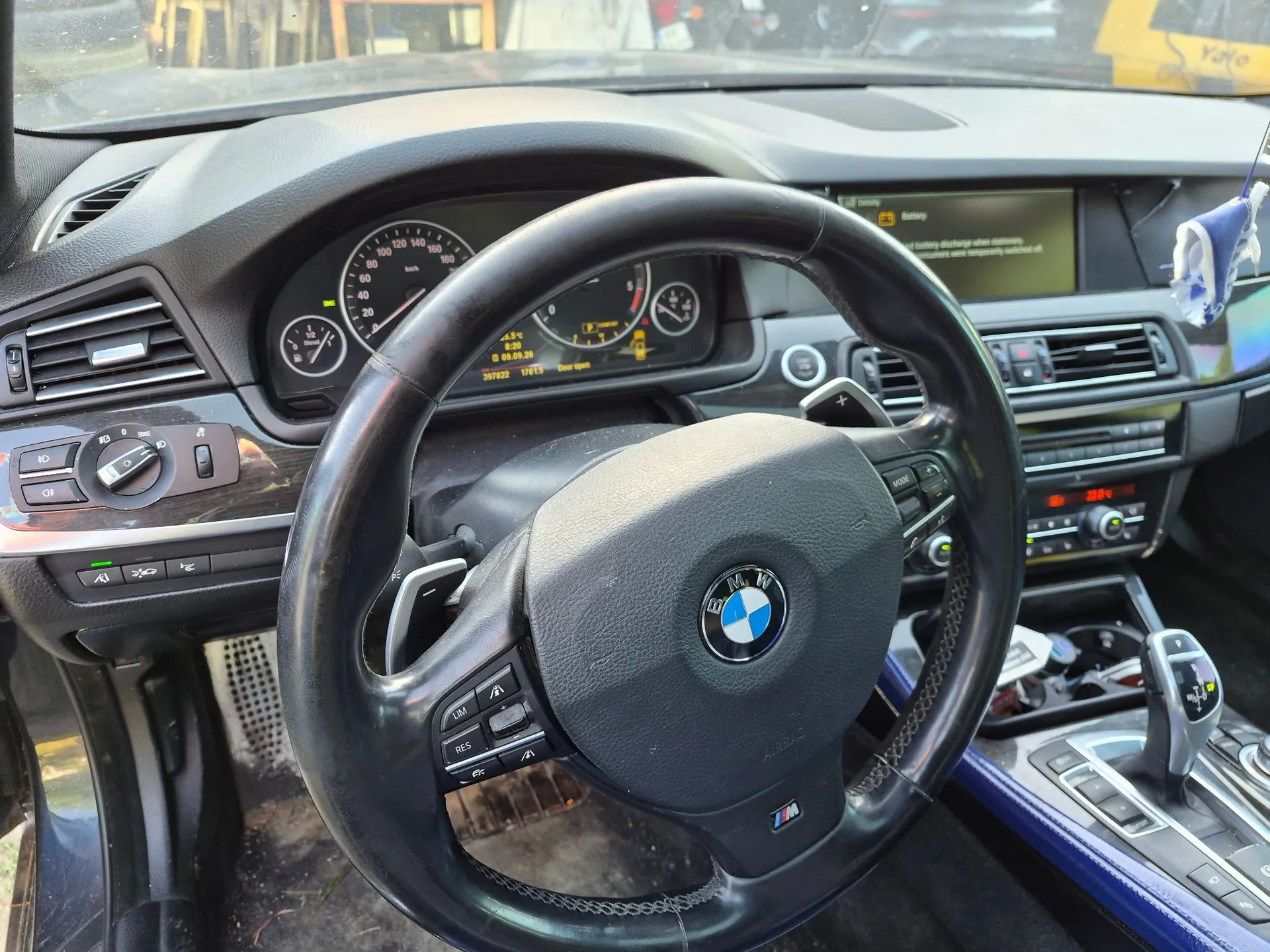 Wnętrze samochodu marki BMW z perspektywy kierowcy. Widoczna jest czarna kierownica z logo BMW w centrum i przyciskami wielofunkcyjnymi, deska rozdzielcza z zegarami analogowymi i cyfrowym wyświetlaczem pokazującym datę 09.05.20, przebieg 207822 km oraz temperaturę -0.5°C. Po prawej stronie znajduje się konsola środkowa z ekranem multimedialnym, nawiewami i lewarkiem zmiany biegów.
