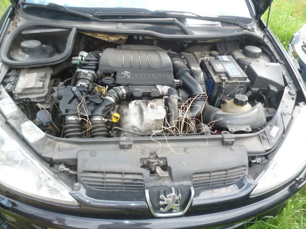Widok z góry na otwartą maskę ciemnego samochodu marki Peugeot, ukazujący silnik spalinowy z napisem HDI na plastikowej osłonie. Widoczny jest akumulator po prawej stronie, układ dolotowy oraz elementy mechaniczne silnika. Na przednim grillu znajduje się srebrne logo lwa Peugeot.