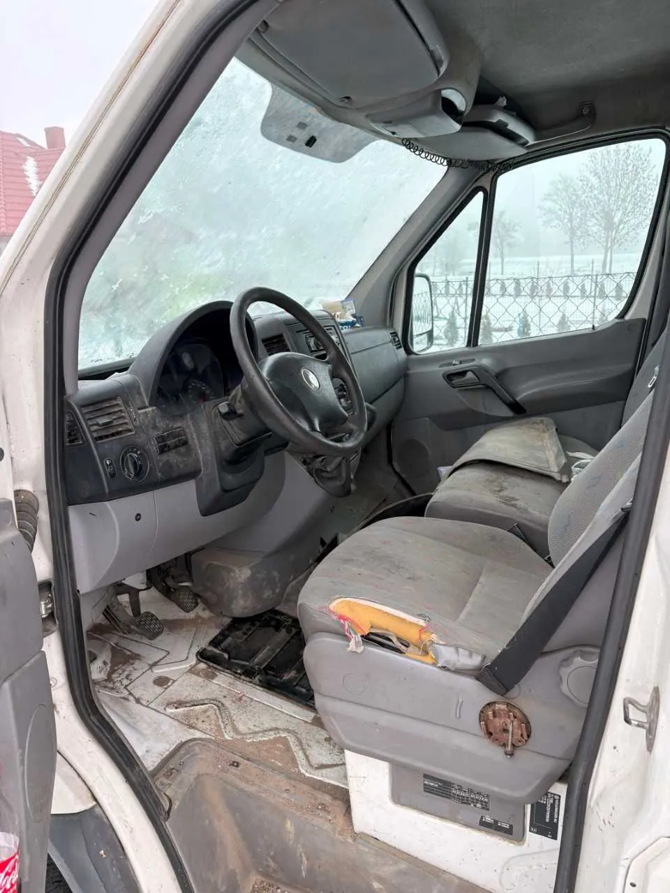 Wnętrze furgonetki z otwartą drzwiami od strony kierowcy. Widoczne są zniszczone, brudne siedzenia w kolorze szarym, z uszkodzeniem na siedzeniu kierowcy (wyciekający żółty materiał). Kierownica z logo w centrum, zniszczona deska rozdzielcza z przyciskami i wentylacją. Na podłodze brudne dywaniki i odsłonięte elementy podłogi. Po lewej stronie widoczna butelka Coca-Coli. Przez przednią szybę widać zimowy krajobraz z opadającym śniegiem, ogrodzeniem i drzewami. Zewnętrzne tło obejmuje czerwony dach budynku i pochmurne niebo.