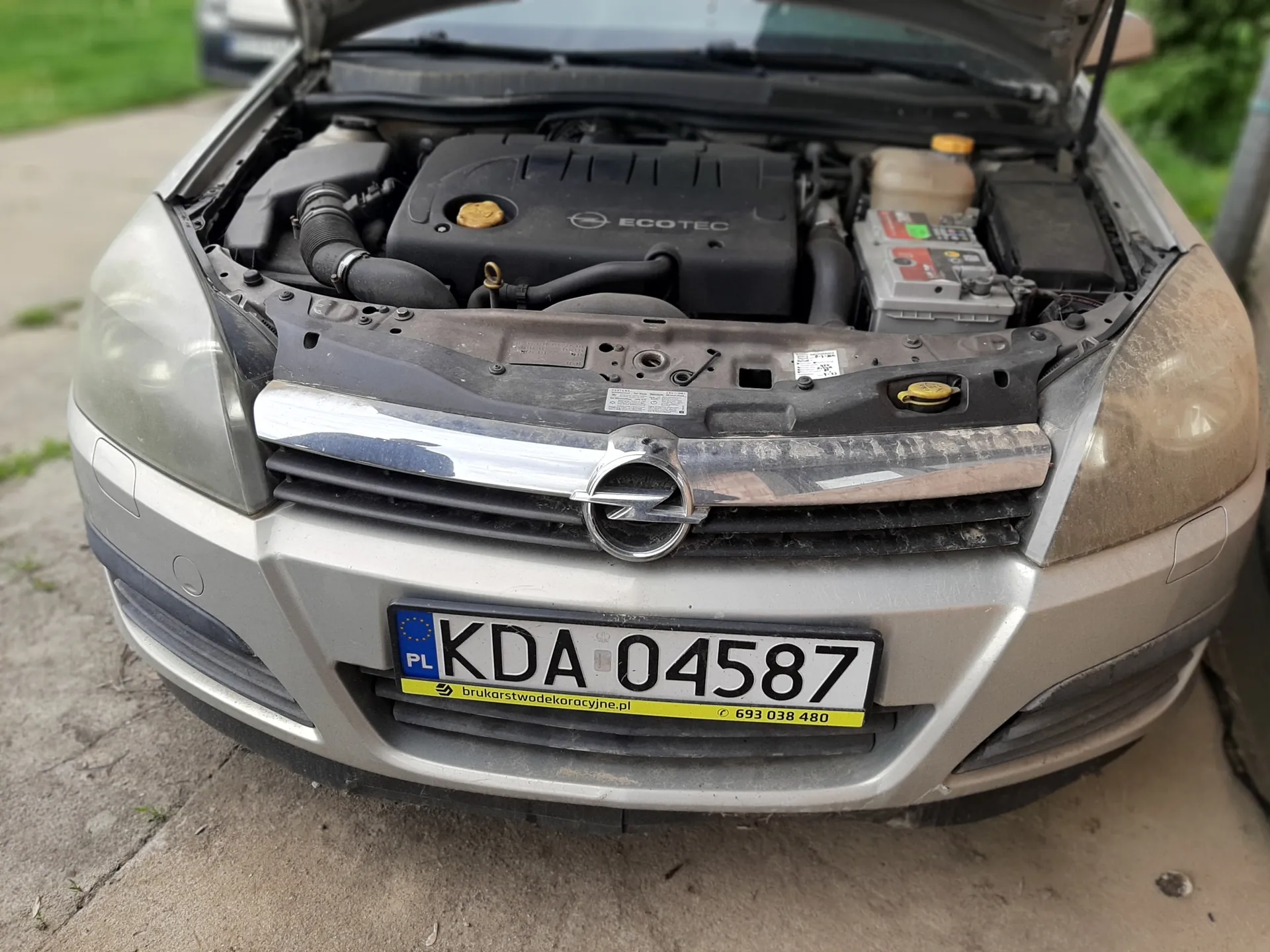 Przednia część srebrnego samochodu marki Opel z otwartą klapą silnika. Widoczny silnik z napisem ECOTEC, tablica rejestracyjna KDA 04587 z niebieskim pasem PL po lewej stronie oraz żółtą naklejką z tekstem brukarstwodekoracyjne.pl i numerem telefonu 693 038 480. Samochód stoi na betonowej powierzchni, w tle zielona trawa i fragment innego pojazdu.