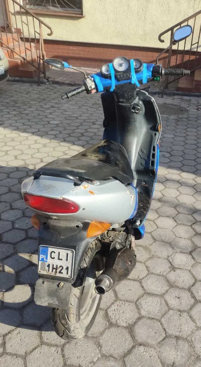 Scooter o kolorze niebiesko-czarnym z srebrnym tyłem, zaparkowany na chodniku z sześciokątnych płyt. Widoczna tablica rejestracyjna PL CLI 1H21, niebieska taśma na kierownicy, lusterka boczne oraz tylne światło. W tle budynek z ceglaną podmurówką, schodami i oknem.