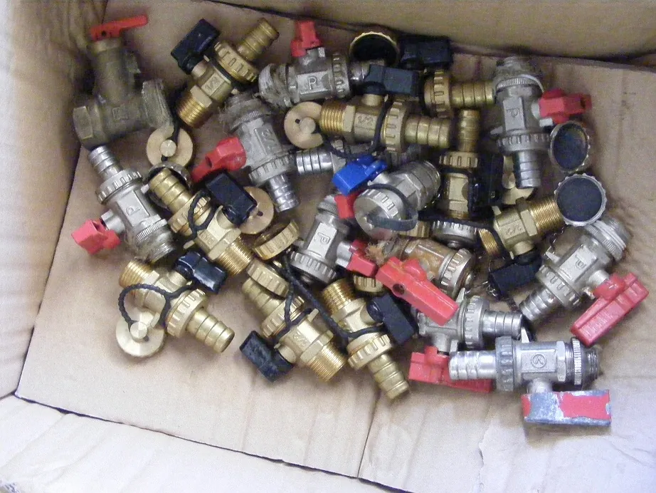 Kartonowe pudełko wypełnione licznymi metalowymi zaworami i złączkami hydraulicznymi. Elementy są wykonane z mosiądzu i stali, niektóre posiadają czerwone lub niebieskie plastikowe uchwyty oraz czarne nakrętki. Część elementów jest powiązana czarnymi gumkami lub sznurkami.