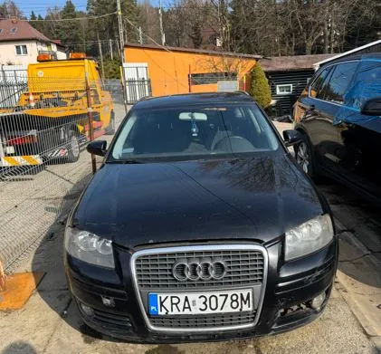 Czarny samochód marki Audi z tablicą rejestracyjną KRA-3078M na pierwszym planie. Po prawej stronie widoczny jest czarny samochód, a w tle żółty pojazd specjalistyczny, pomarańczowy budynek, drzewa i ogrodzenie. Scena rozgrywa się na otwartym placu z betonowym podłożem.