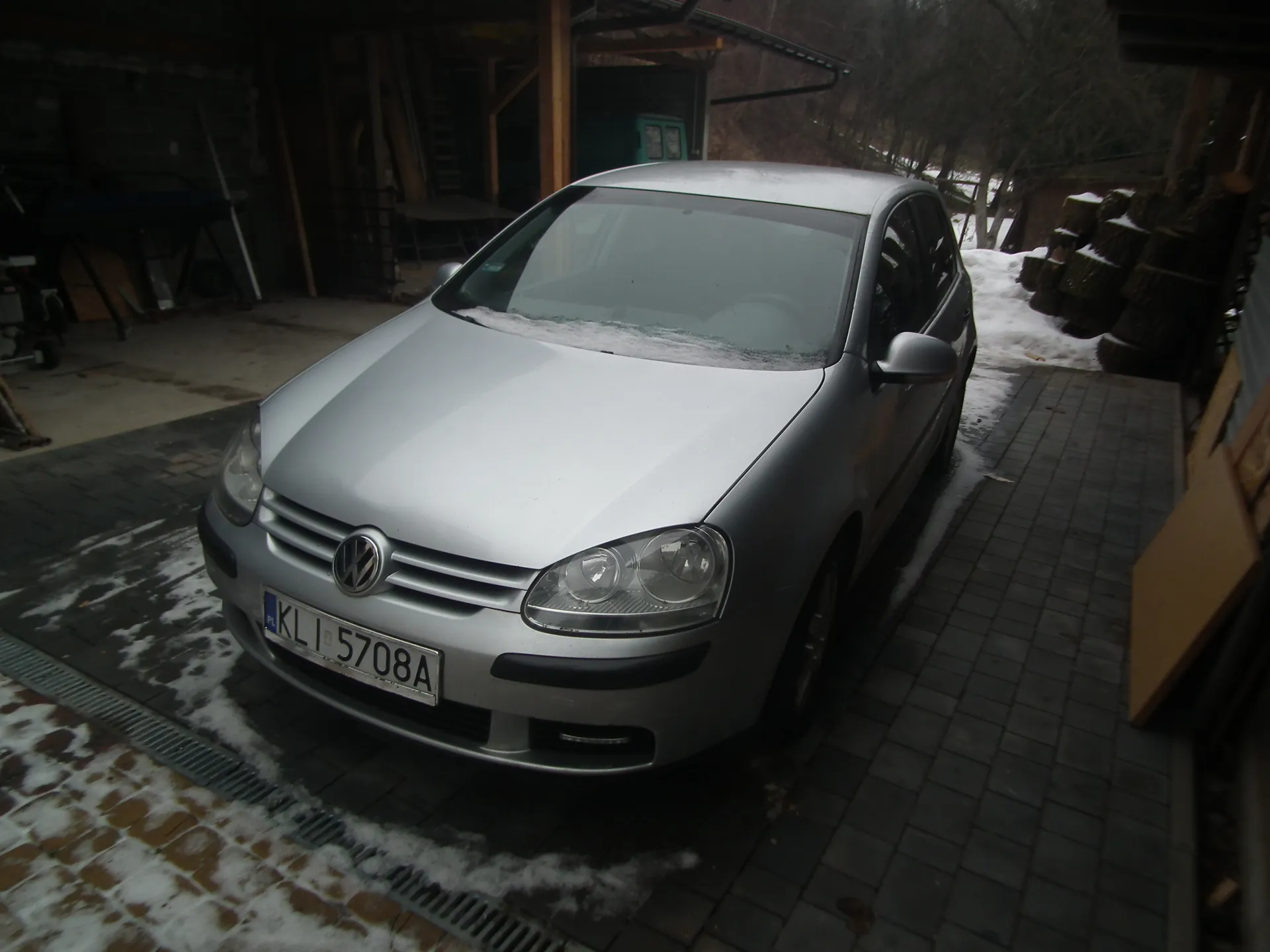 Srebrny samochód marki Volkswagen Golf z tablicą rejestracyjną KLI 5708A zaparkowany na brukowanej powierzchni. Na masce i ziemi widoczny śnieg. W tle budynek garażowy z narzędziami, stosami drewna i drzewami bez liści. Horyzont i tekst na tablicy są poziome, obraz jest prawidłowo zorientowany.
