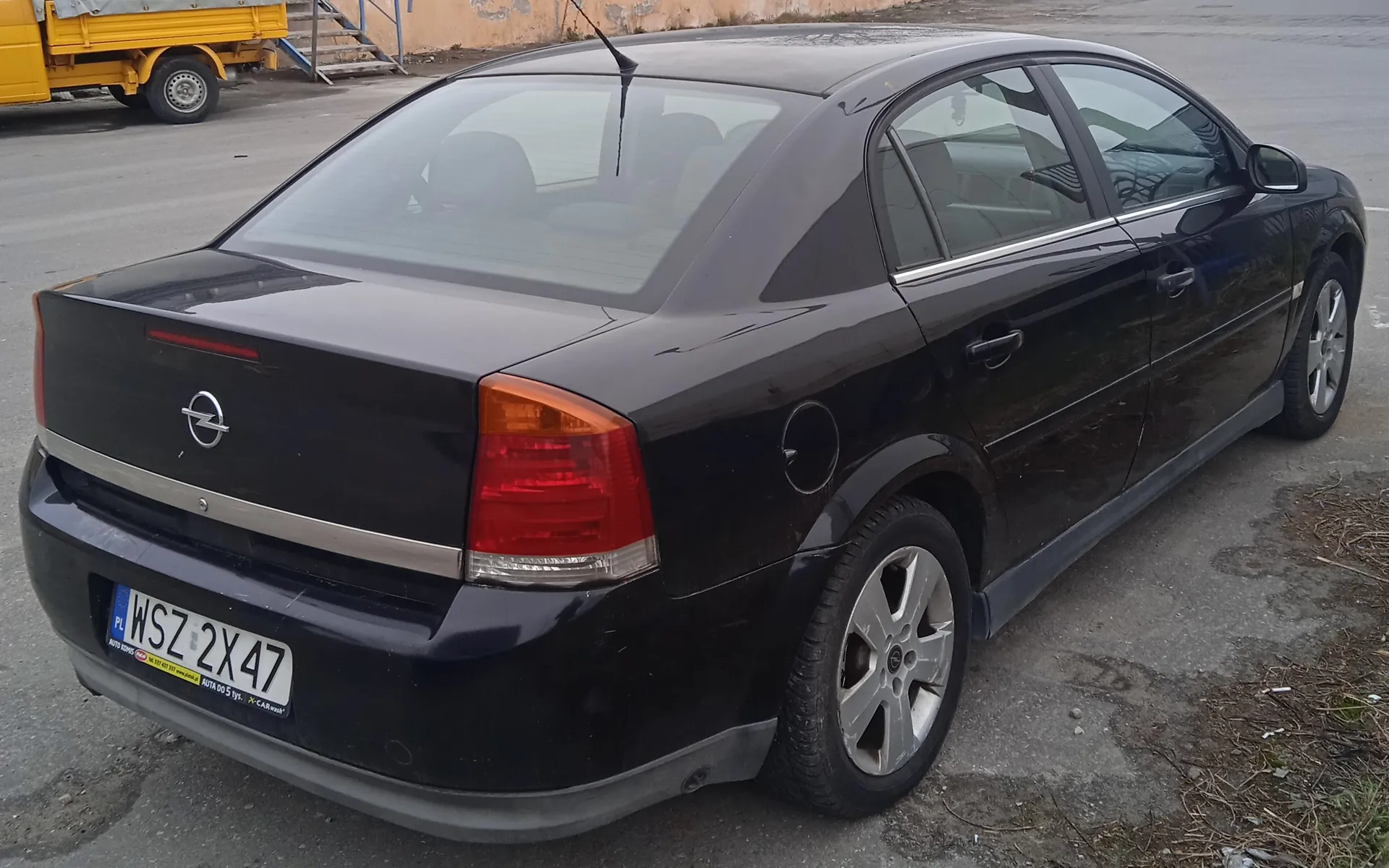 Czarny samochód osobowy marki Opel, widok z tyłu i boku. Tablica rejestracyjna z napisem WSZ 2X47 oraz oznaczeniem PL. W tle żółty samochód ciężarowy, betonowe schody i zniszczona ściana budynku. Samochód zaparkowany na asfaltowej powierzchni z widocznymi plamami i suchą trawą.