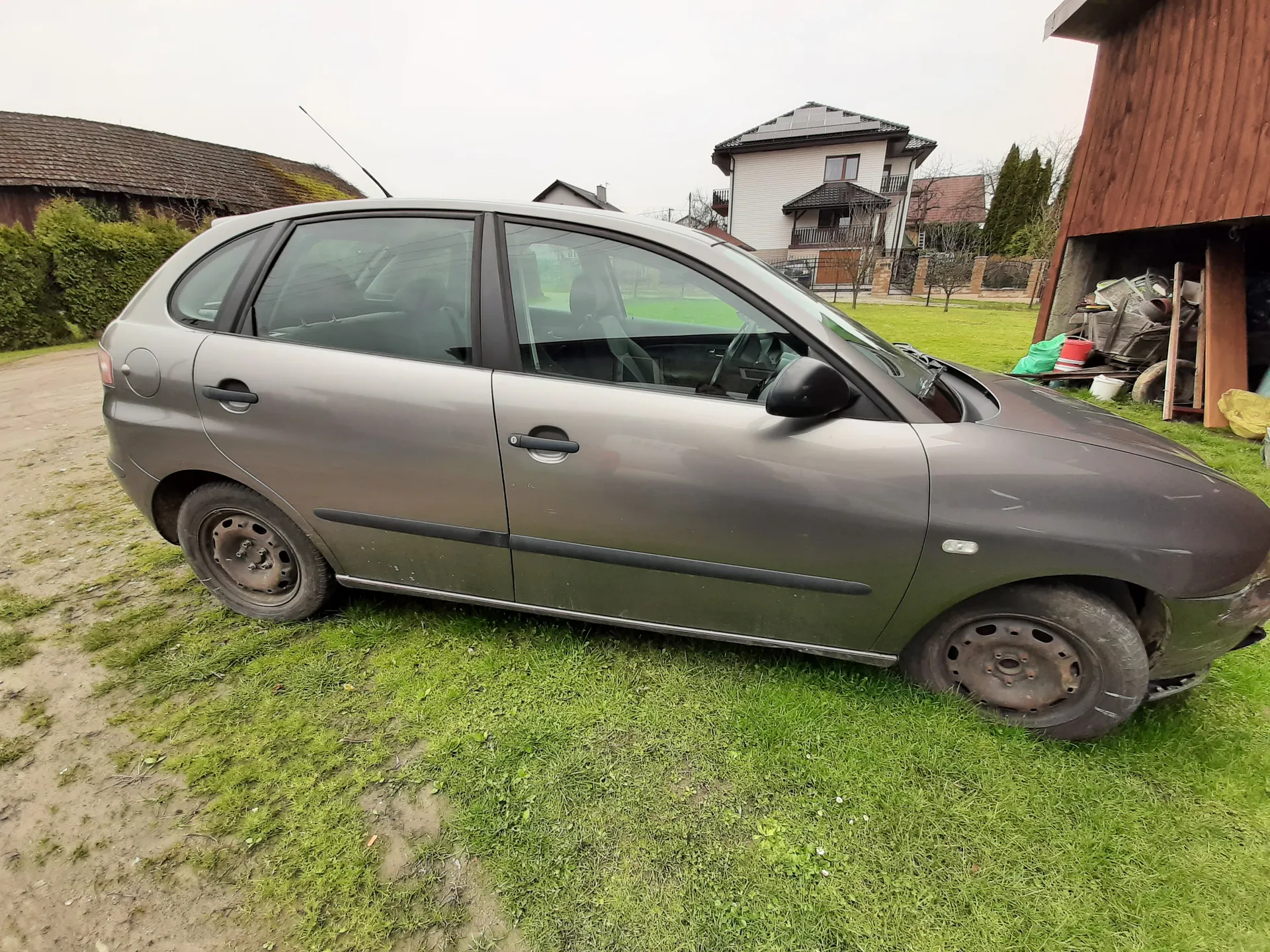 Szary samochód osobowy typu hatchback zaparkowany na trawie, z widocznymi oznakami zużycia i brakiem opony na przednim prawym kole. W tle domy mieszkalne, zielona trawa, brudna droga oraz drewniany garaż z narzędziami i pojemnikami. Niebo jest pochmurne.