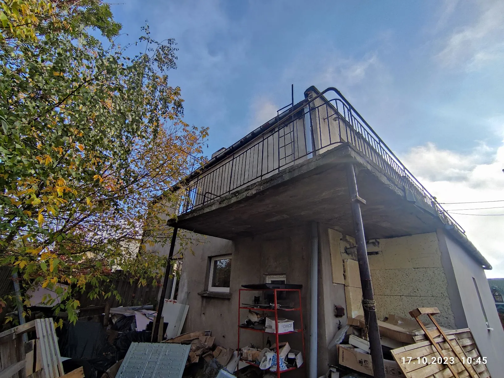 Zdjęcie przedstawiające fragment szarego, betonowego budynku z balkonem na piętrze, otoczonym metalową balustradą. Pod balkonem znajduje się czerwony regał z różnymi przedmiotami oraz sterta materiałów budowlanych i odpadów. Po lewej stronie widoczne jest drzewo z żółknącymi liśćmi. W prawym dolnym rogu znajduje się data i godzina: 17.10.2023 10:45.