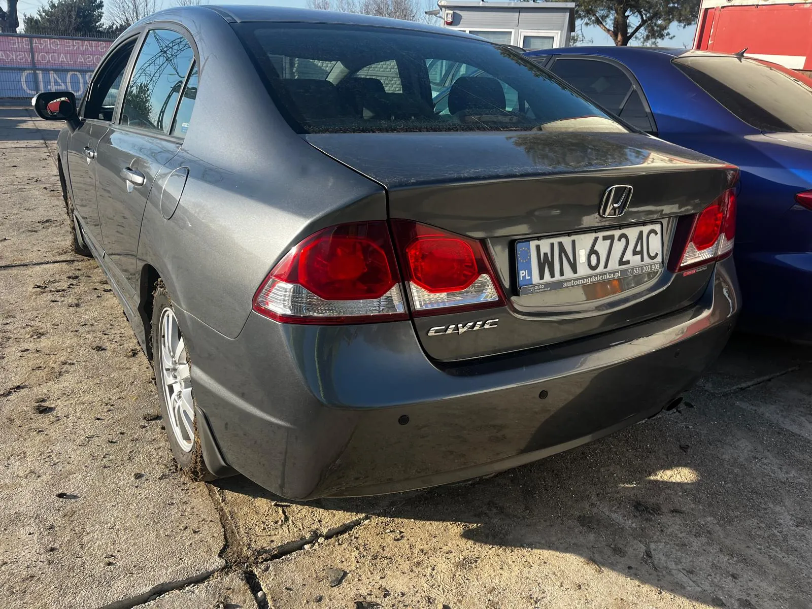 Tył szarego samochodu Honda Civic z tablicą rejestracyjną WN 6724C (PL), widocznym logo Hondy i napisem "CIVIC". Na tablicy dodatkowo widoczny adres strony internetowej automagdalenka.pl oraz numer telefonu 531 202 505. Samochód zaparkowany na betonowej powierzchni z pęknięciami, obok niego niebieski samochód. W tle budynek, drzewa i fragment ogrodzenia z częściowo widocznym napisem "CIAO".
