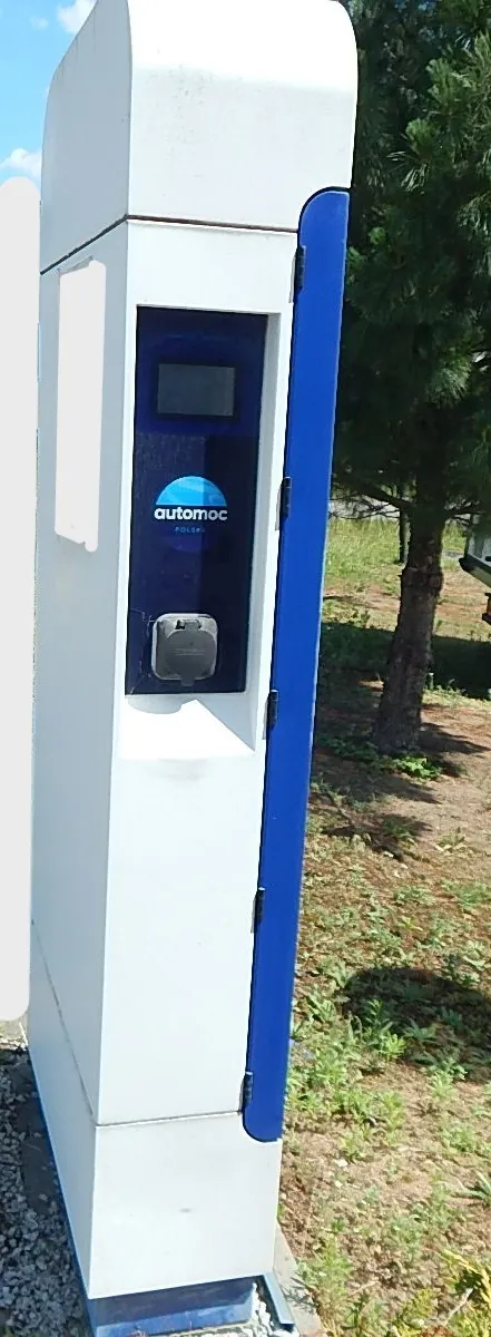Biały, pionowy słup stacji ładowania pojazdów elektrycznych z niebieskim panelem bocznym. Na przednim panelu znajduje się ekran, logo 'automoc POLSKA' oraz gniazdo ładowania. Urządzenie stoi na zewnątrz, w tle widoczna jest trawa i drzewo iglaste.