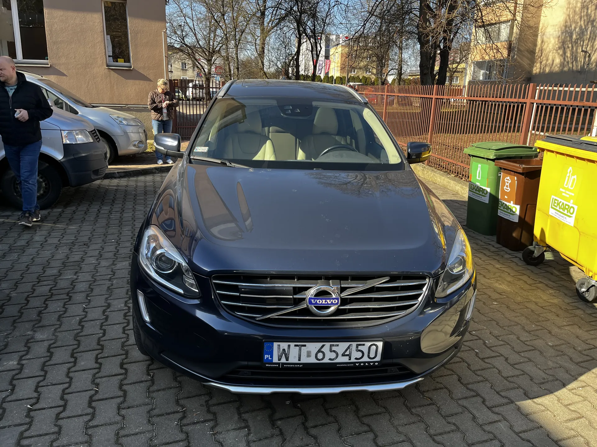 Ciemnoniebieski samochód marki Volvo z tablicą rejestracyjną PL WT 65450 zaparkowany na brukowanej powierzchni. Przed samochodem widoczne są trzy pojemniki na śmieci: zielony, brązowy i żółty z napisem EKARO. W tle budynki, ogrodzenie, drzewa bez liści oraz dwie osoby: mężczyzna w czarnej kurtce i niebieskich spodniach idący po lewej stronie oraz osoba w brązowej kurtce stojąca blisko ogrodzenia. Na masce samochodu znajduje się logo VOLVO.