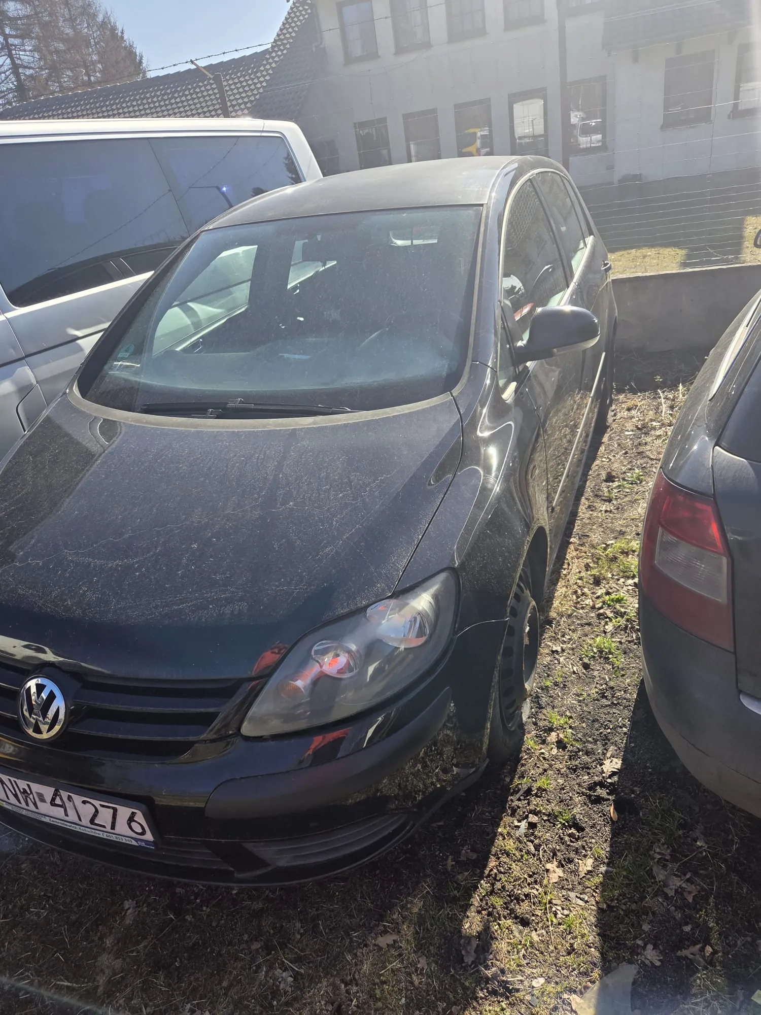 Czarny samochód marki Volkswagen z tablicą rejestracyjną NH 41276 zaparkowany na trawiastym terenie. W tle widoczne są inne pojazdy, budynek z oknami oraz ogrodzenie. Samochód pokryty jest kurzem, a na przedniej szybie widać ślady. Tło obejmuje również drzewa i jasne niebo.