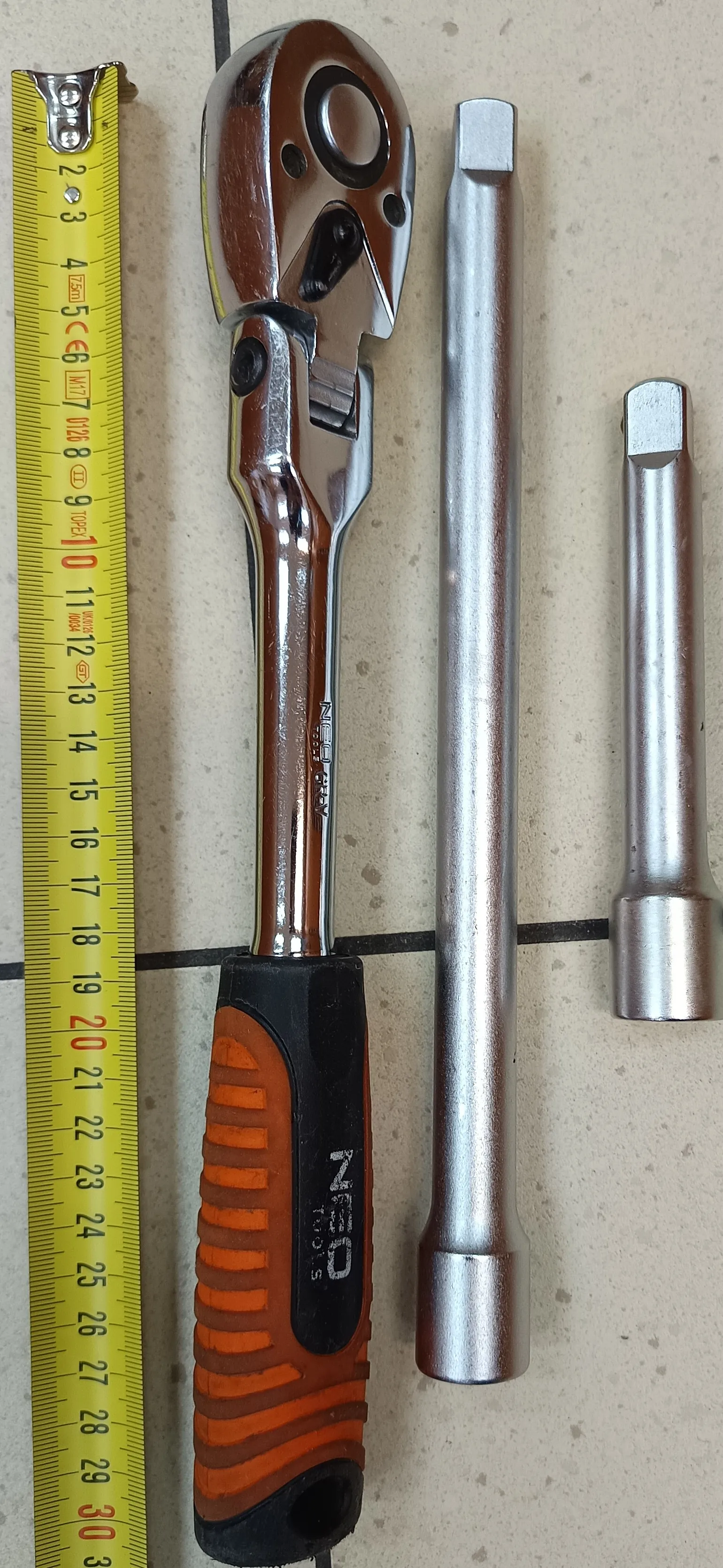 Na jasnym tle leżą cztery przedmioty: żółta taśma miernicza z czarnymi i czerwonymi oznaczeniami (liczby od 2 do 30, widoczne napisy CE, M7), klucz ręczny z chromowanym ciałem i pomarańczowo-czarną rączką z napisem NEO, dwa metalowe nasadki (dłuższa i krótsza) o srebrnym kolorze. Wszystkie przedmioty ułożone są pionowo.